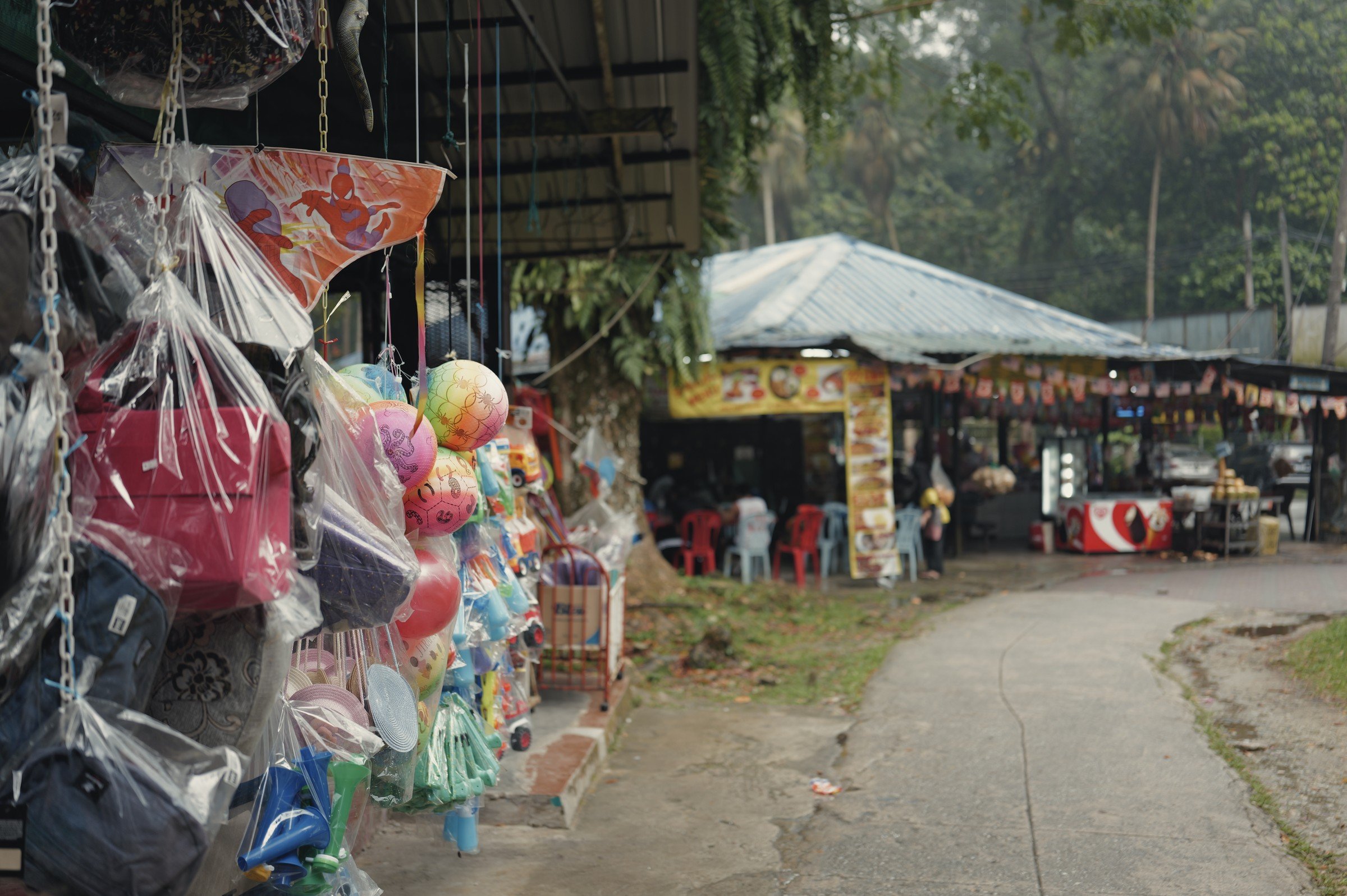 Colourful toys hang beside small stalls on a tree-lined path.