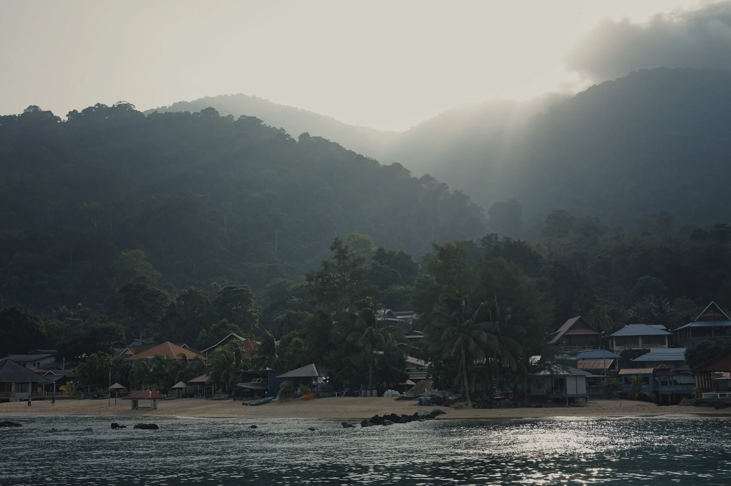 A serene coastal village with houses along the shoreline, backed by lush, forested mountains shrouded in mist, and a partly cloudy sky.