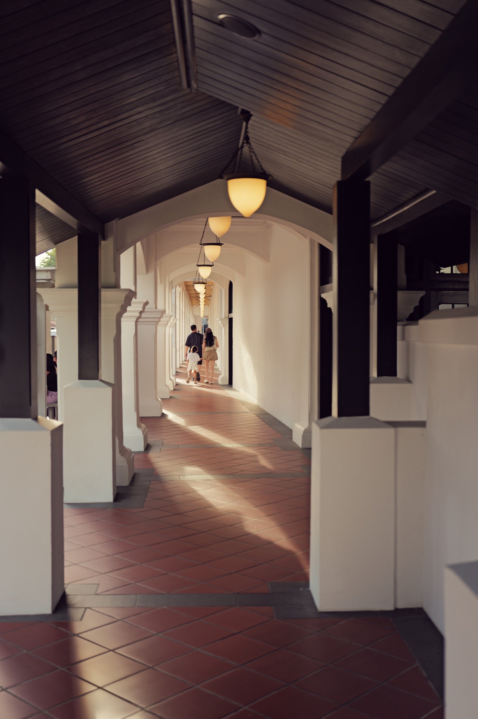 A row of white columns and warm ceiling lamps recedes along a sheltered tiled walkway.