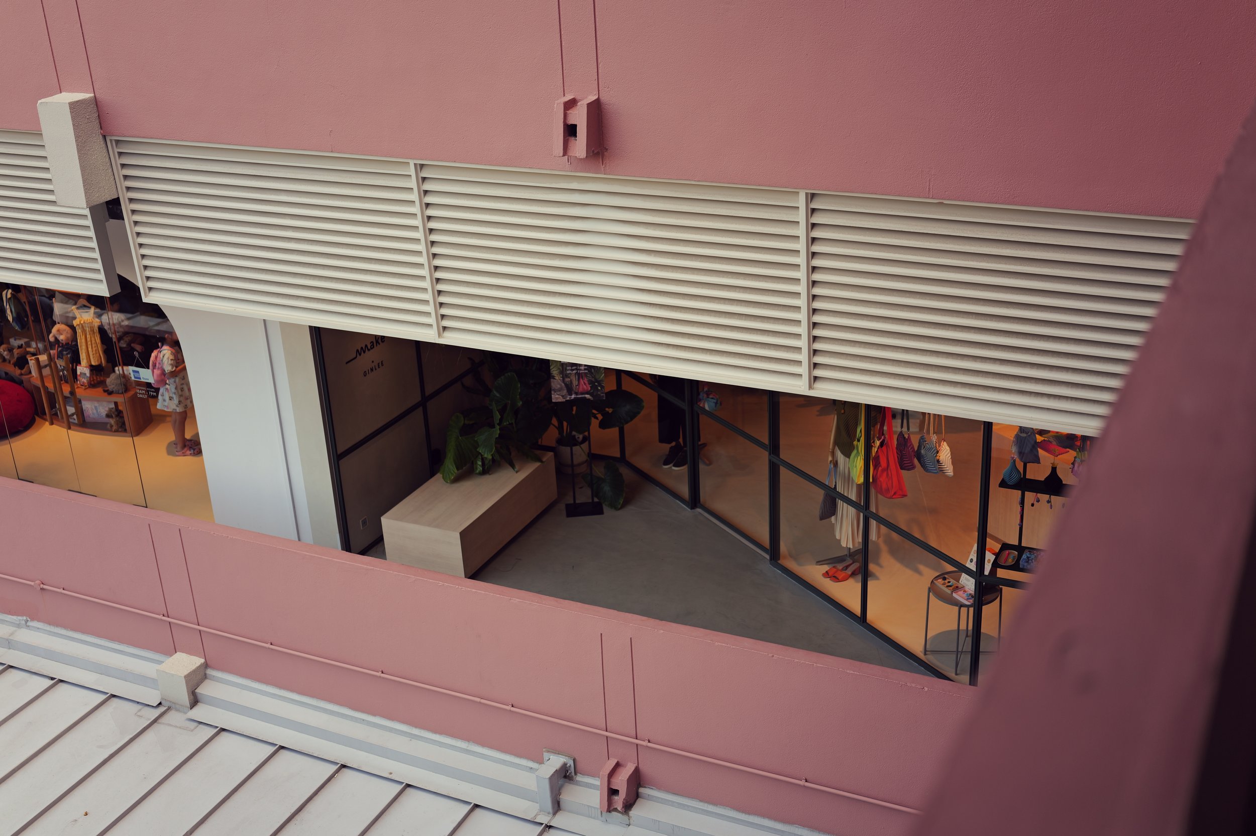 View of a shopping mall or retail store from above, with clothing displays and mannequins visible through large glass windows. The exterior wall of the building is painted pink, with metal ventilation grates.