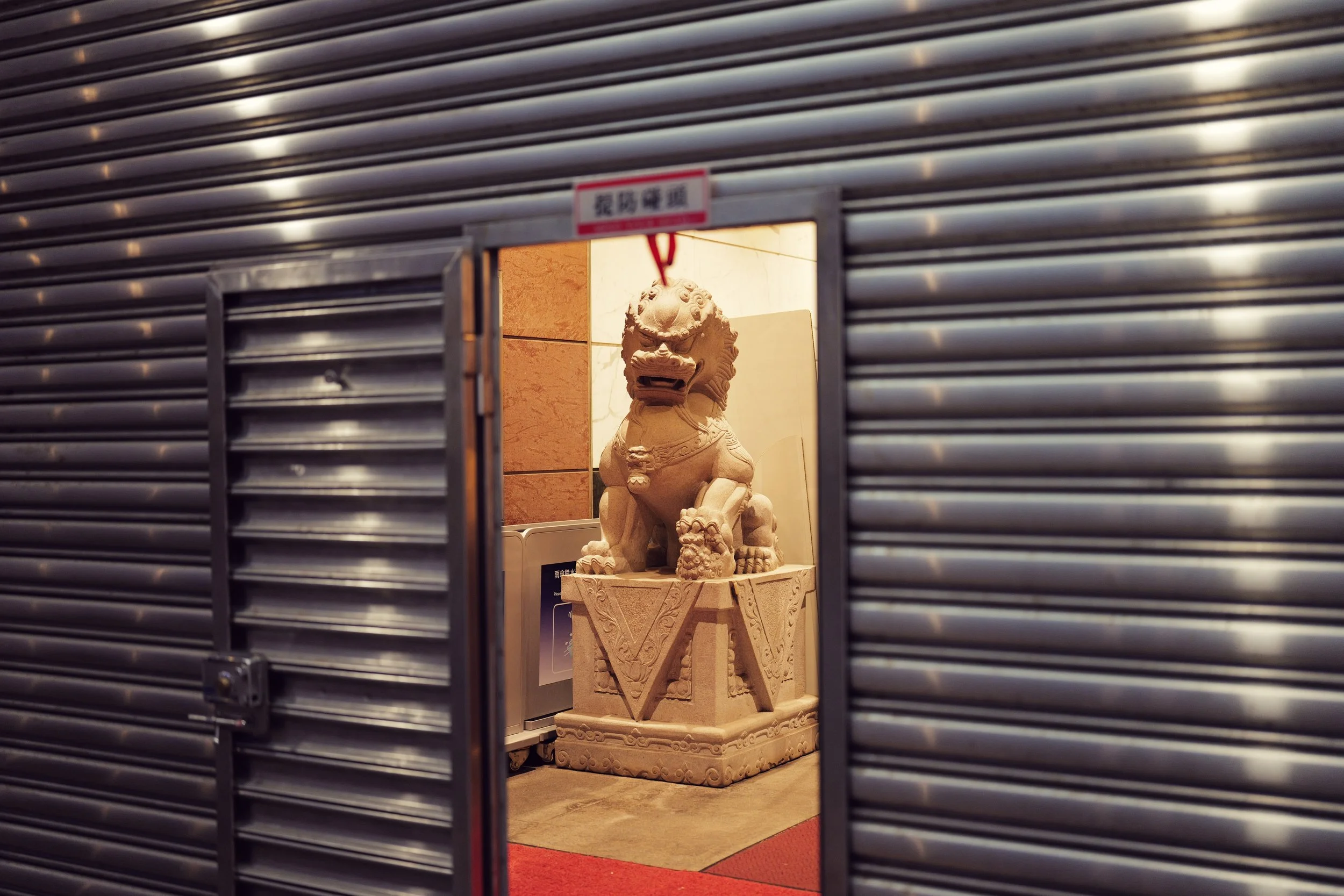 A stone Chinese guardian lion sculpture on a pedestal, seen through a partially opened metal shutter.