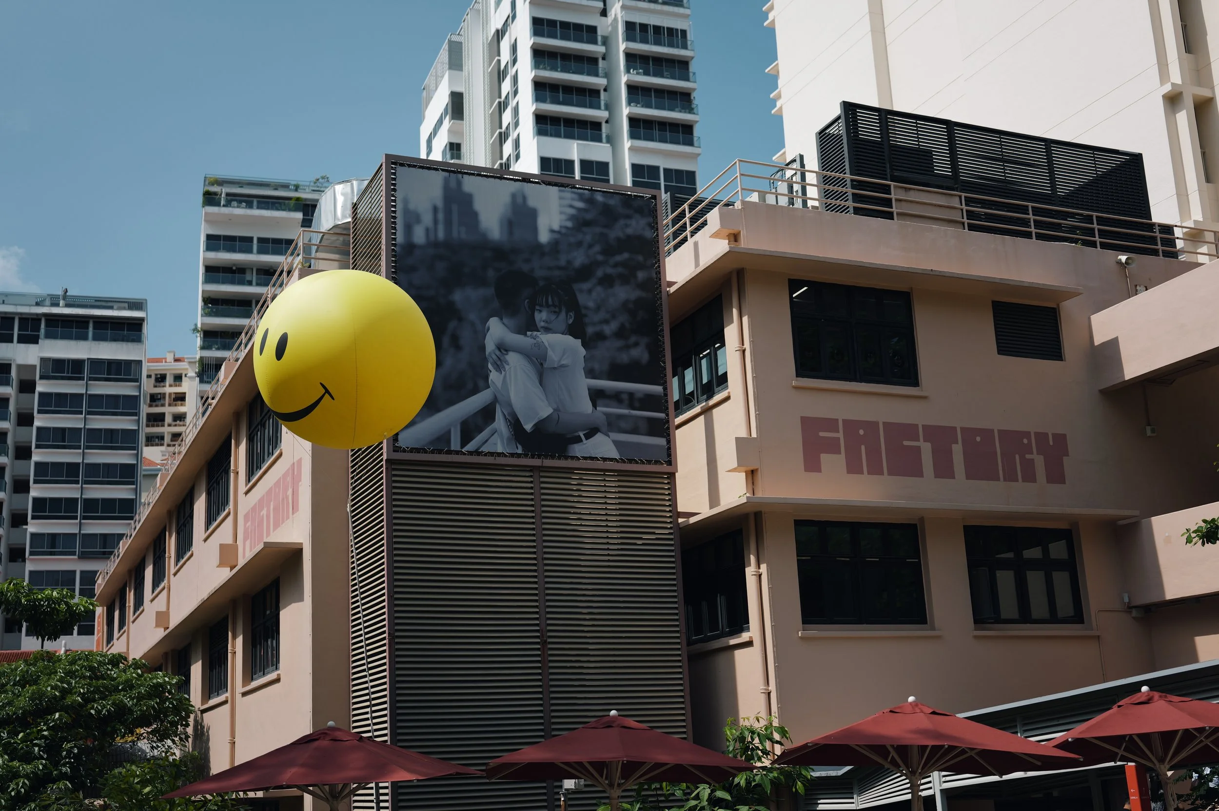 Urban scene with a building featuring large black and white advertising posters, a yellow smiley face balloon, and red umbrellas at the bottom.