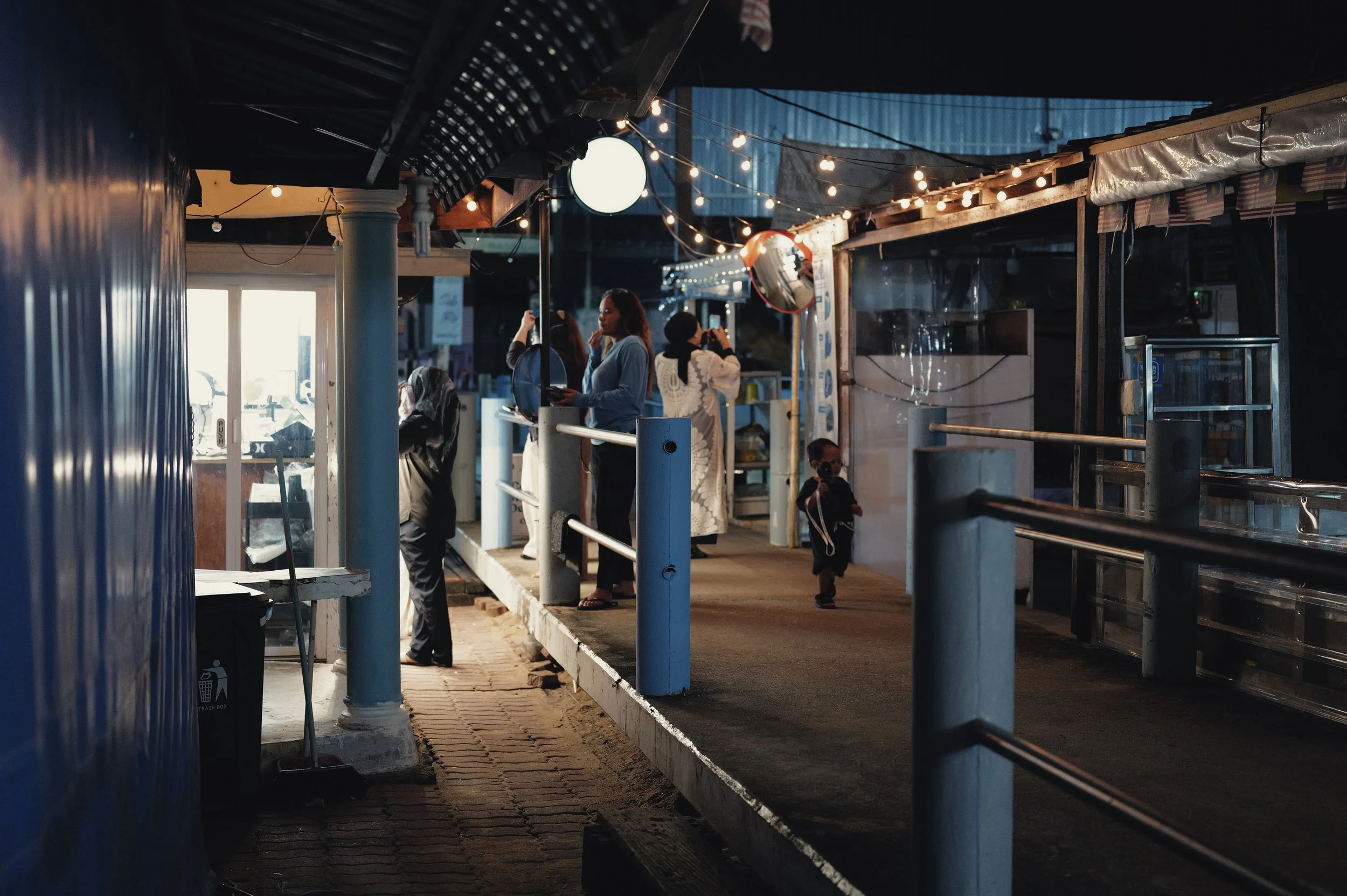 A group of people standing and walking in an outdoor market or food stall area during evening time, with string lights hanging overhead and a blue building in the background.