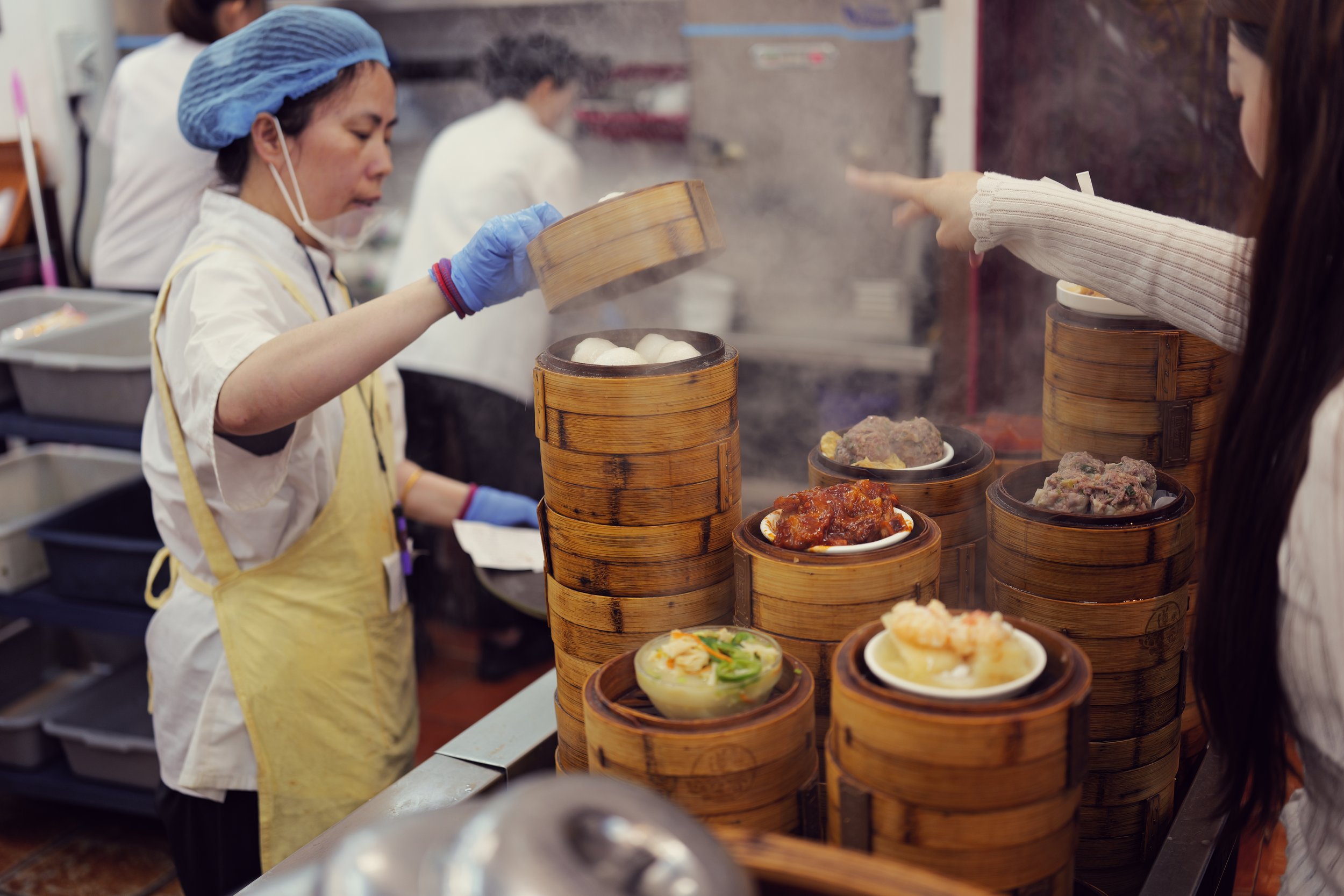 A woman in a yellow apron and blue hairnet serving steamed buns at a food stall, with various dim sum dishes displayed in bamboo steamers.