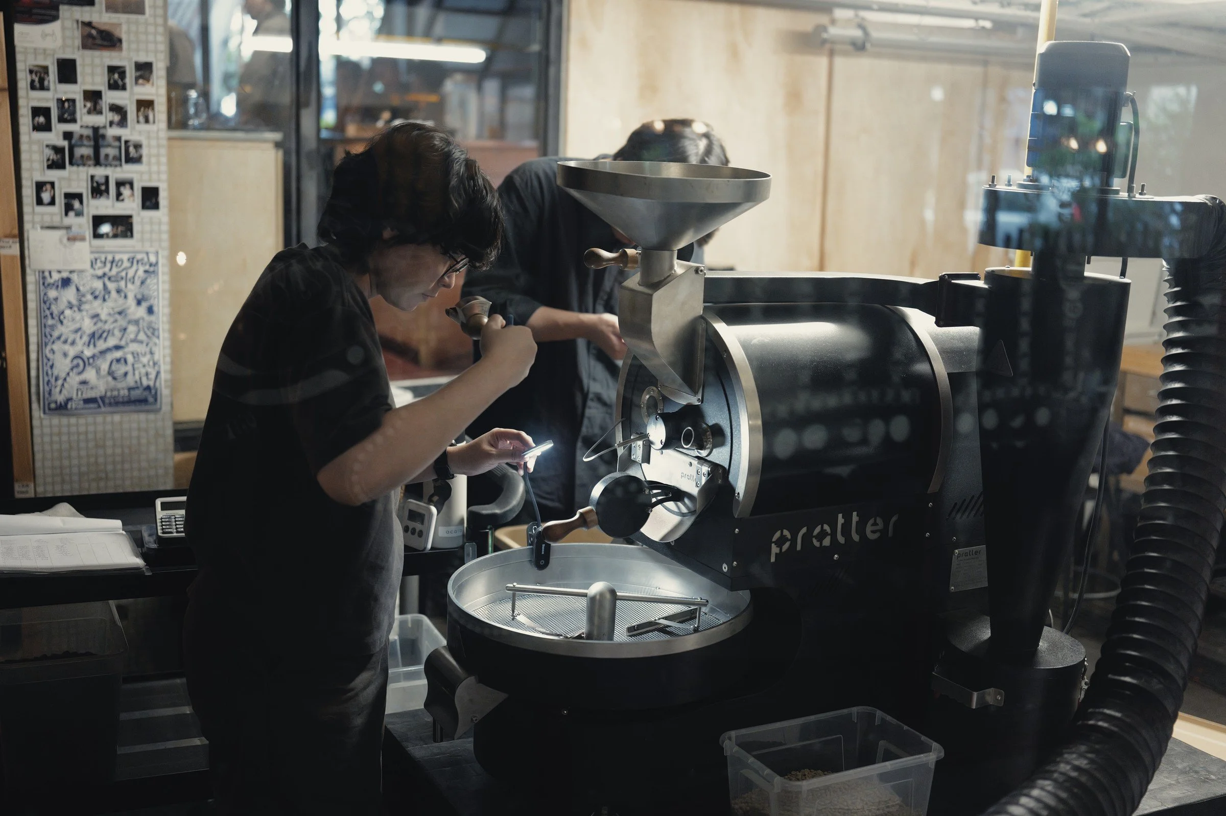 A worker leans over a coffee-roasting machine beside a window.