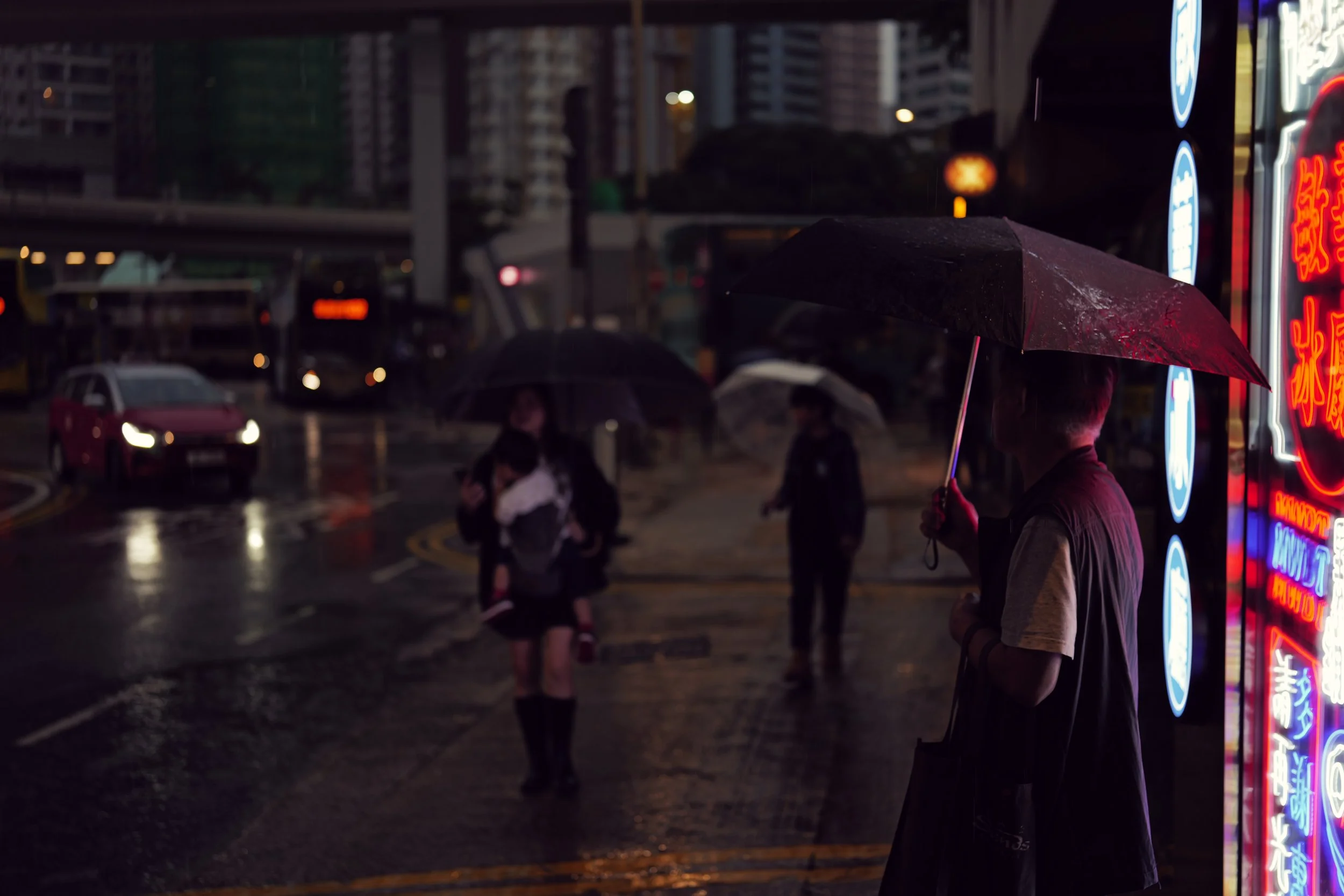 Night scene of a city street with pedestrians holding umbrellas, some neon signs visible and buildings in the background.