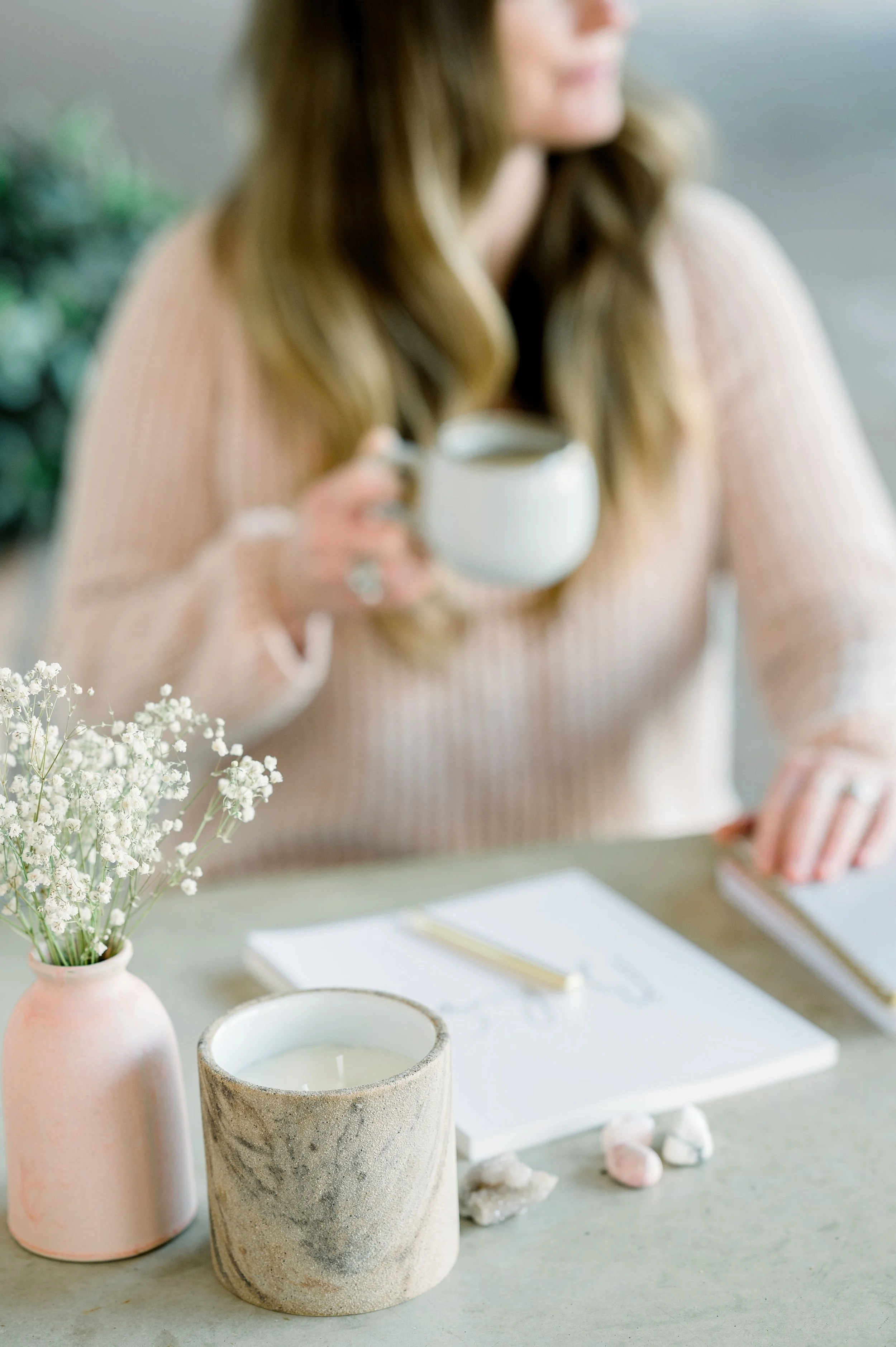 A woman holding a white mug, with decorative items and plants on a table in front of her.