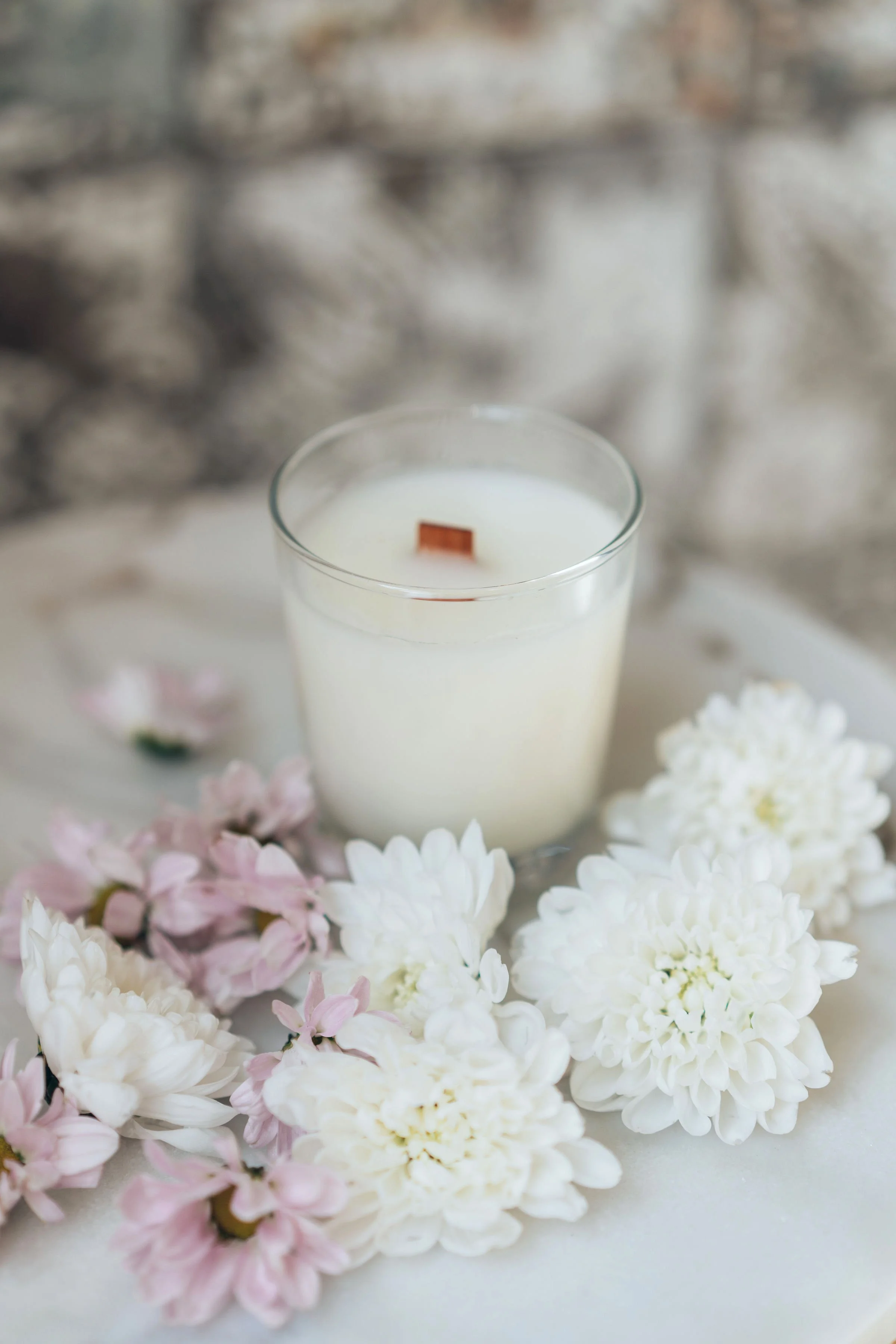 A lit white candle in a clear glass holder surrounded by pink and white flowers against a blurred brick background.