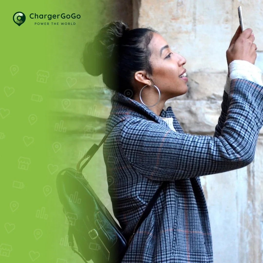 Woman with dark hair in a ponytail wearing hoop earrings, taking a selfie with a smartphone, carrying a black handbag, standing outdoors against a stone wall, with a green overlay on the left side containing company branding and icons.