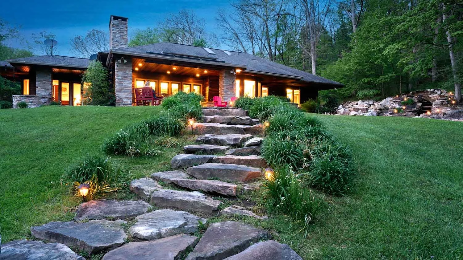 Luxury house with stone steps leading to porch, surrounded by landscaped yard with grass and plants, outdoor lighting, and a waterfall feature in the background, during evening.