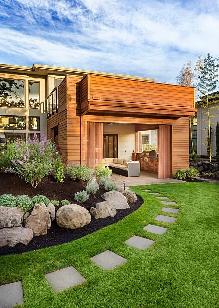 Modern house with wooden exterior and outdoor patio area, situated in a landscaped yard with green lawn, stone walkway, shrubs, and flowering plants.