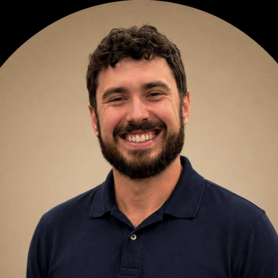 A man with curly brown hair and a beard smiling, wearing a navy blue polo shirt, against a beige background with a black semi-circle at the top