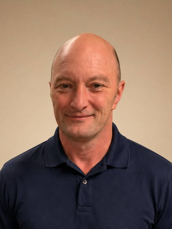 A middle-aged man with a bald head, slight smile, wearing a navy blue collared shirt, standing against a plain beige wall.