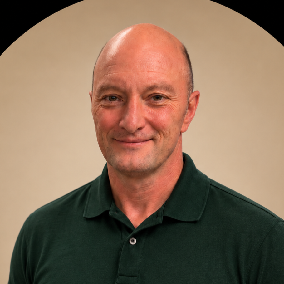 A middle-aged man with a bald head, slight smile, wearing a navy blue collared shirt, standing against a plain beige wall.