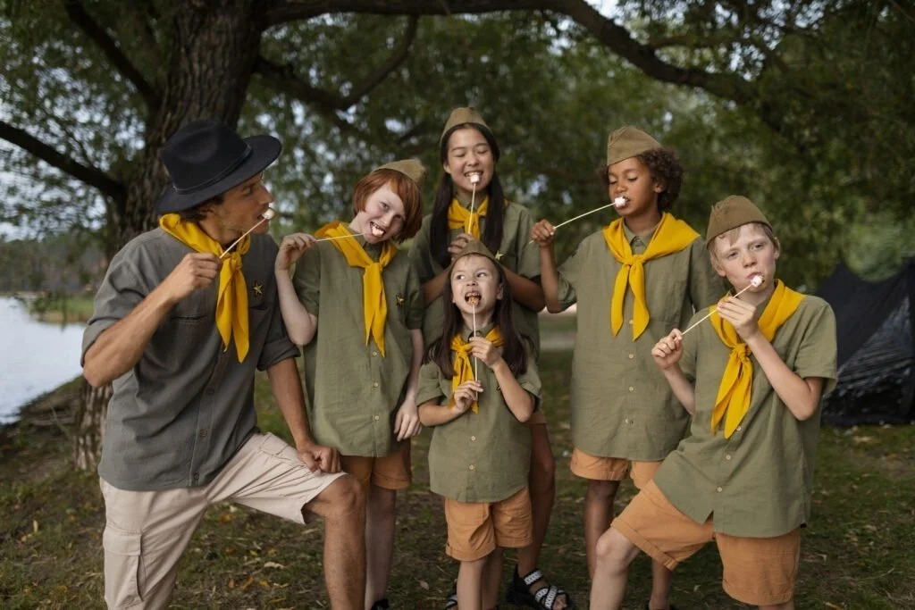 Grupo de campistas niños y un adulto con uniformes y bufandas amarillas haciendo que comen malvaviscos en un ambiente de campamento junto a un río y árboles.