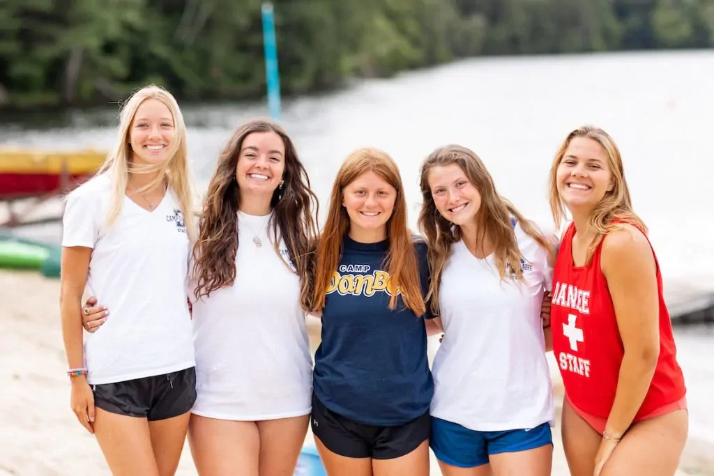 Grupo de cinco chicas sonriendo en la playa al lado de un río, vistiendo ropa de campamento y una de ellas con uniforme de socorrista.