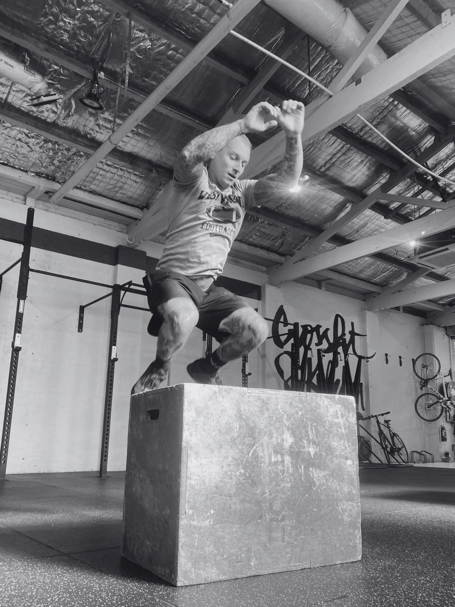 A man performing a box jump onto a sturdy workout box in a gym. The gym has a high ceiling with exposed beams and ventilation pipes, and is equipped with various workout equipment and bicycles.