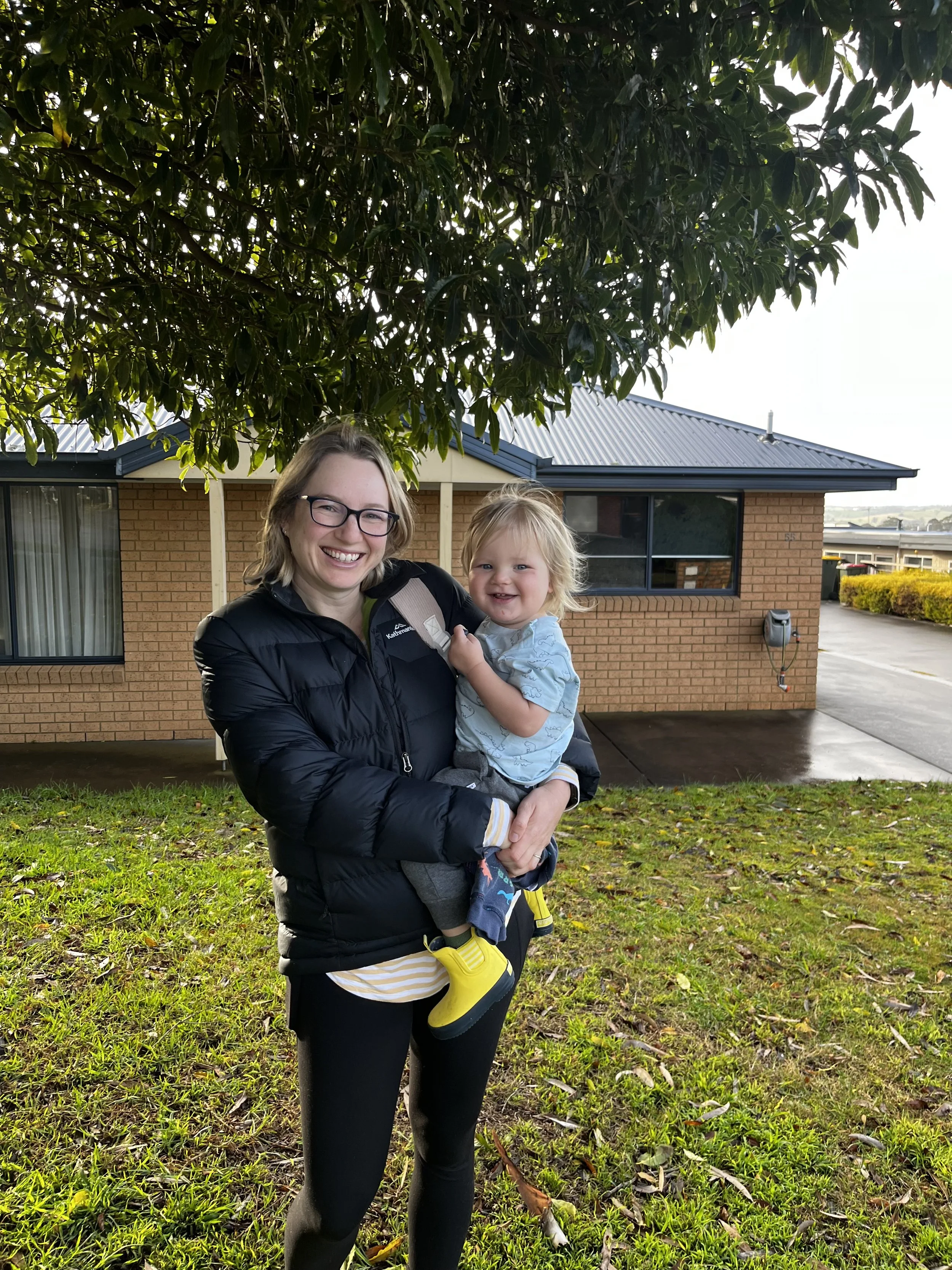 A woman with glasses and a black jacket holding a smiling toddler with blond hair in front of a brick house with a dark roof, a large window, and a small yard with green grass and fallen leaves.