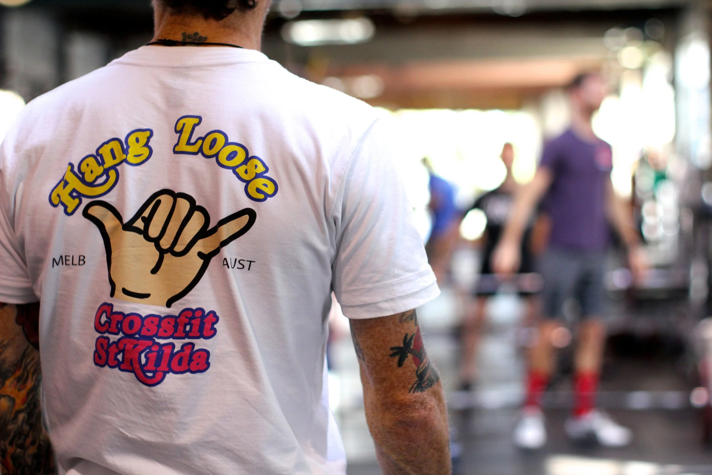 Close-up of a person wearing a white CrossFit T-shirt with colorful text, standing in a gym environment with other people blurred in the background.