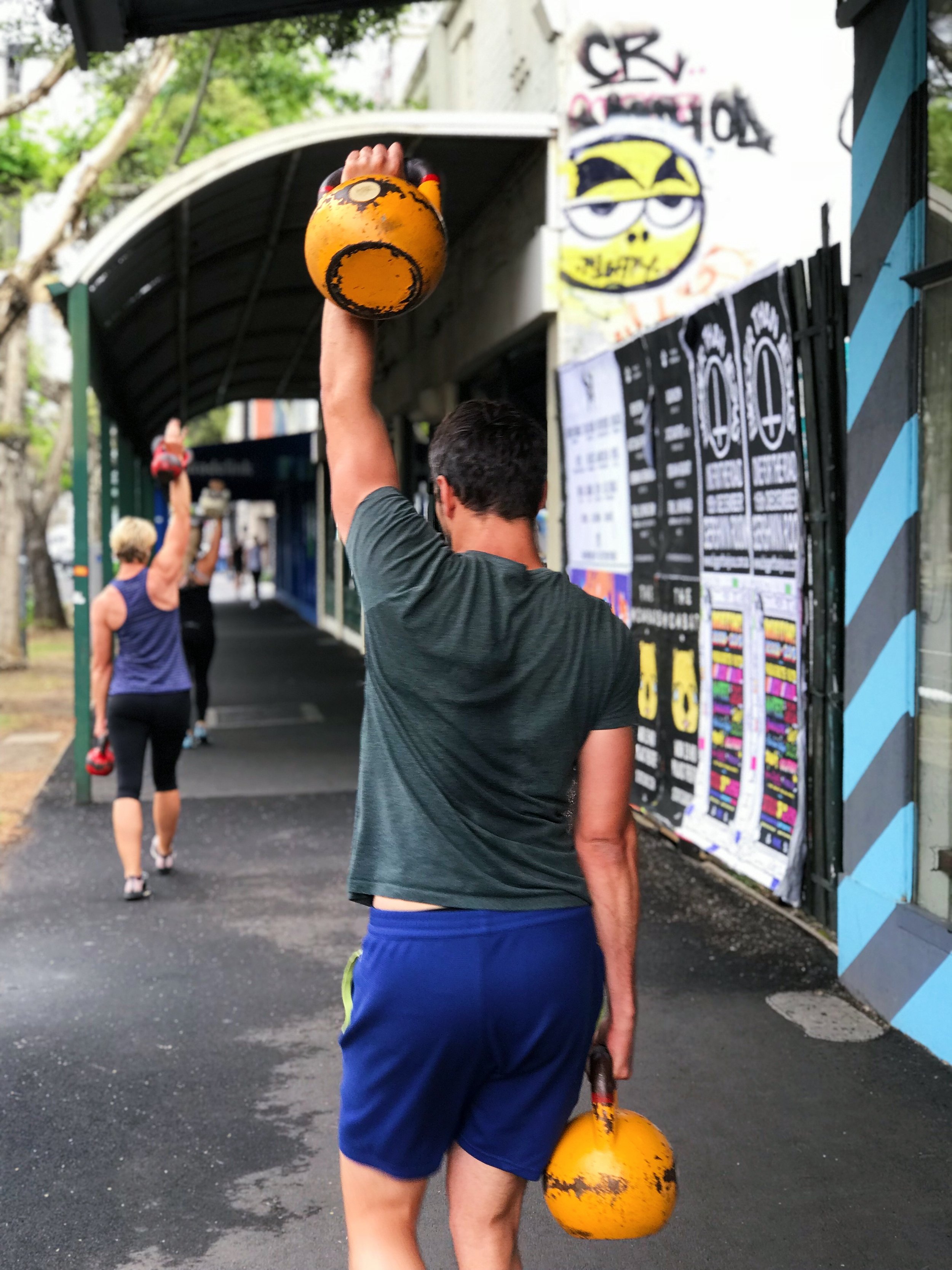 A man walking on a sidewalk holding a yellow kettlebell in his right hand. There are other kettlebells in the background with a covered area and graffiti on the wall.
