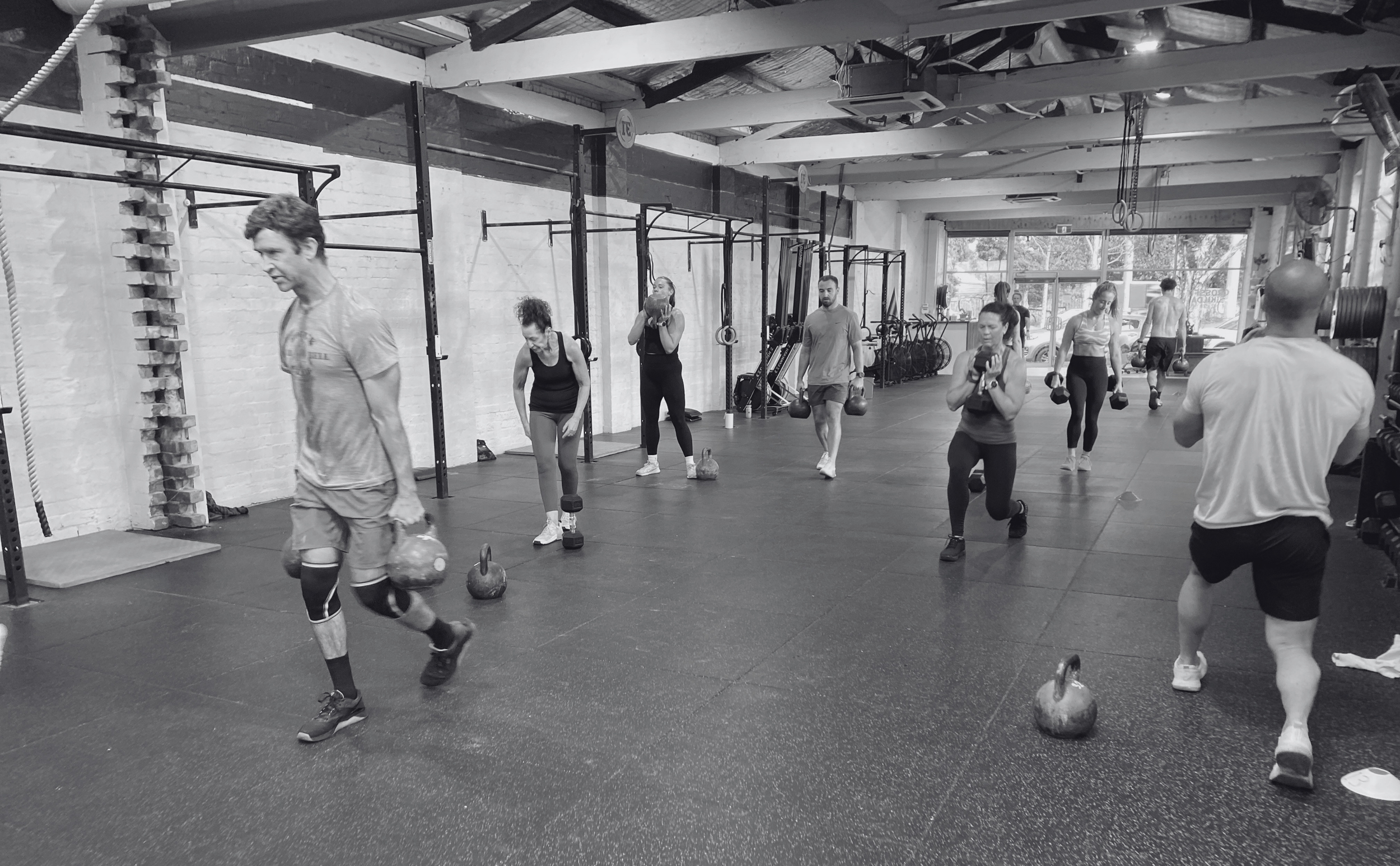 A group of people working out at a gym, lifting kettlebells, and walking around. The gym has high ceilings, large windows, and various fitness equipment.