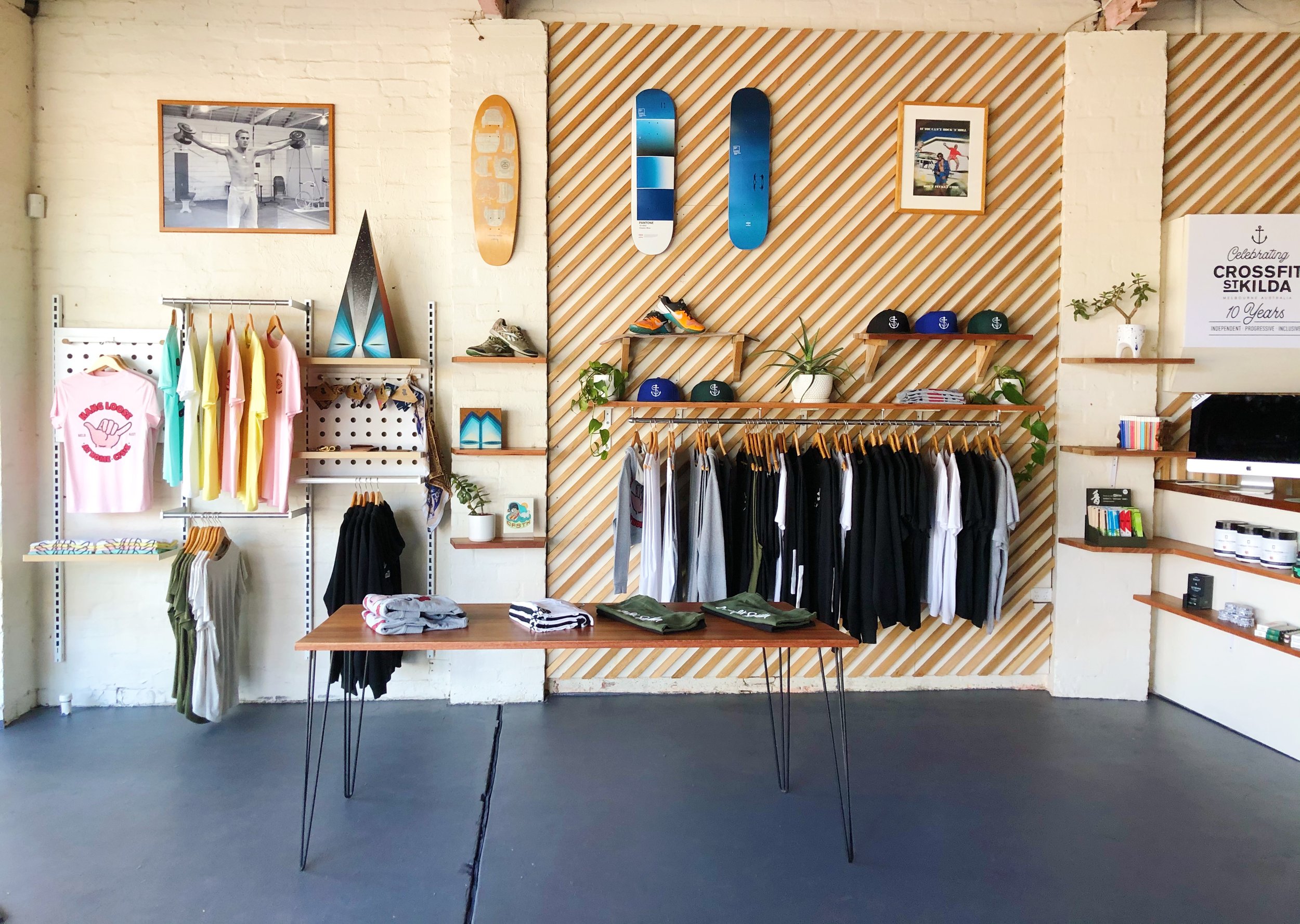Interior view of a retail store with clothing and accessories on display, including t-shirts, hats, skateboards, shoes, and framed photographs on the wall, against a background with a white brick wall and wooden accents.