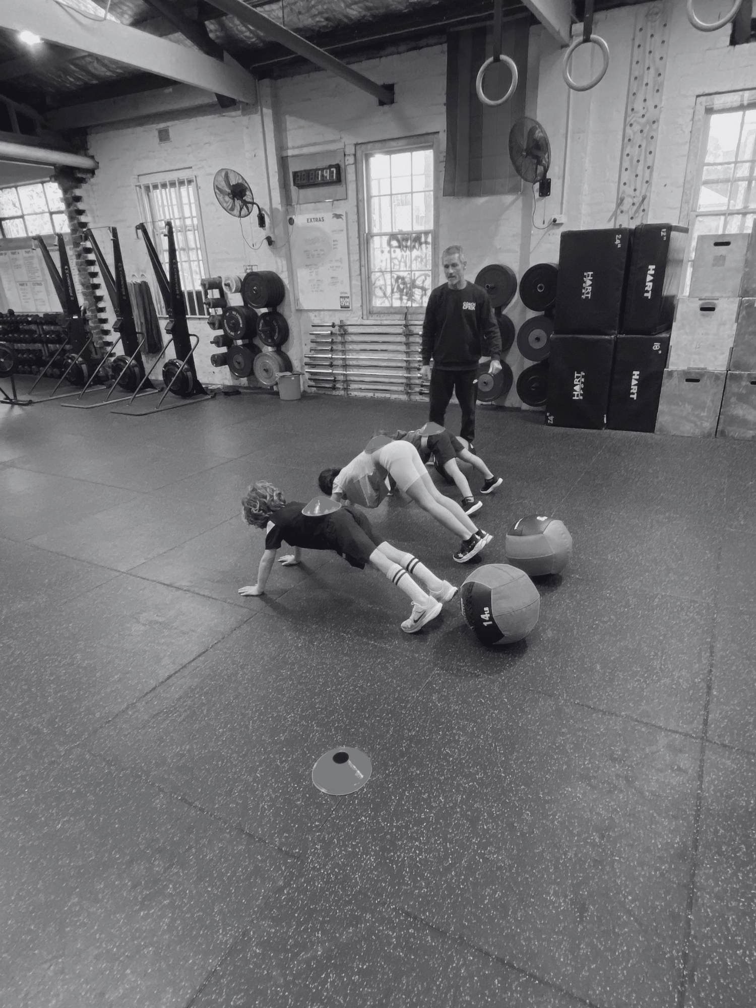 A gym with two children practicing exercises on the floor with a trainer supervising. The gym has various weights and exercise equipment. The image is in black and white.