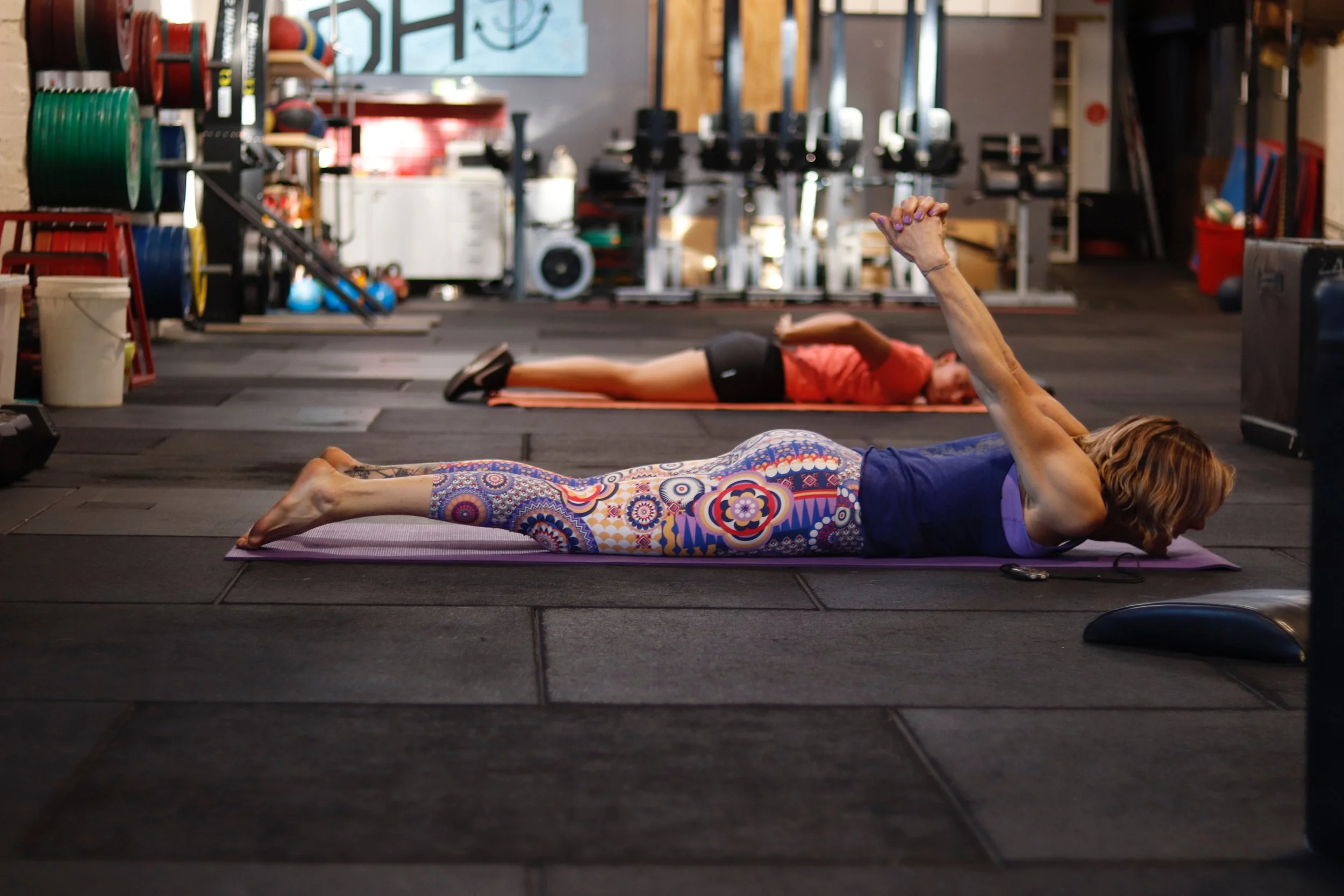 Women doing yoga on mats in a gym with weightlifting equipment in the background.