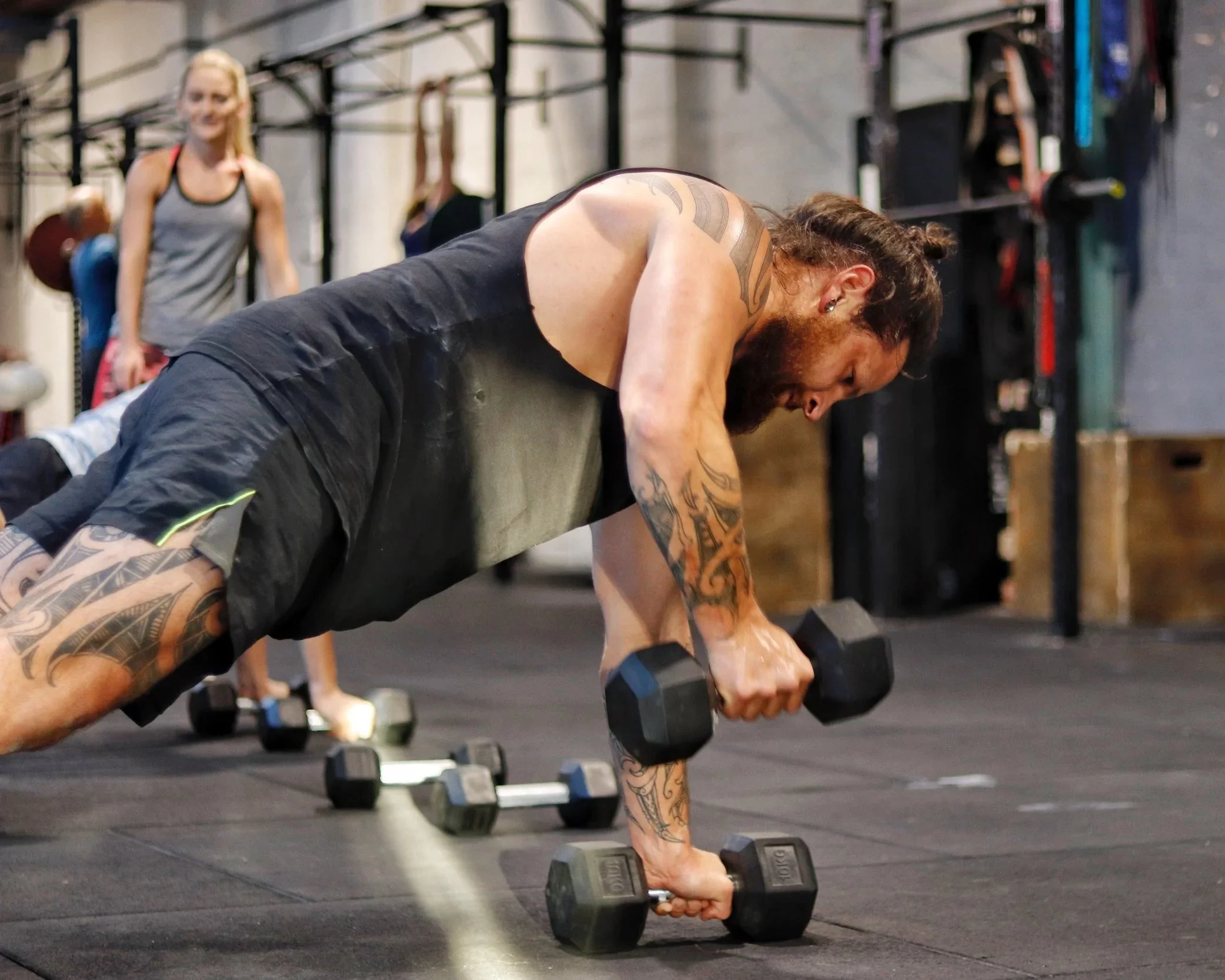 Man with tattoos performing push-ups with dumbbells on the floor of a gym, woman with blonde hair and tank top standing in the background.