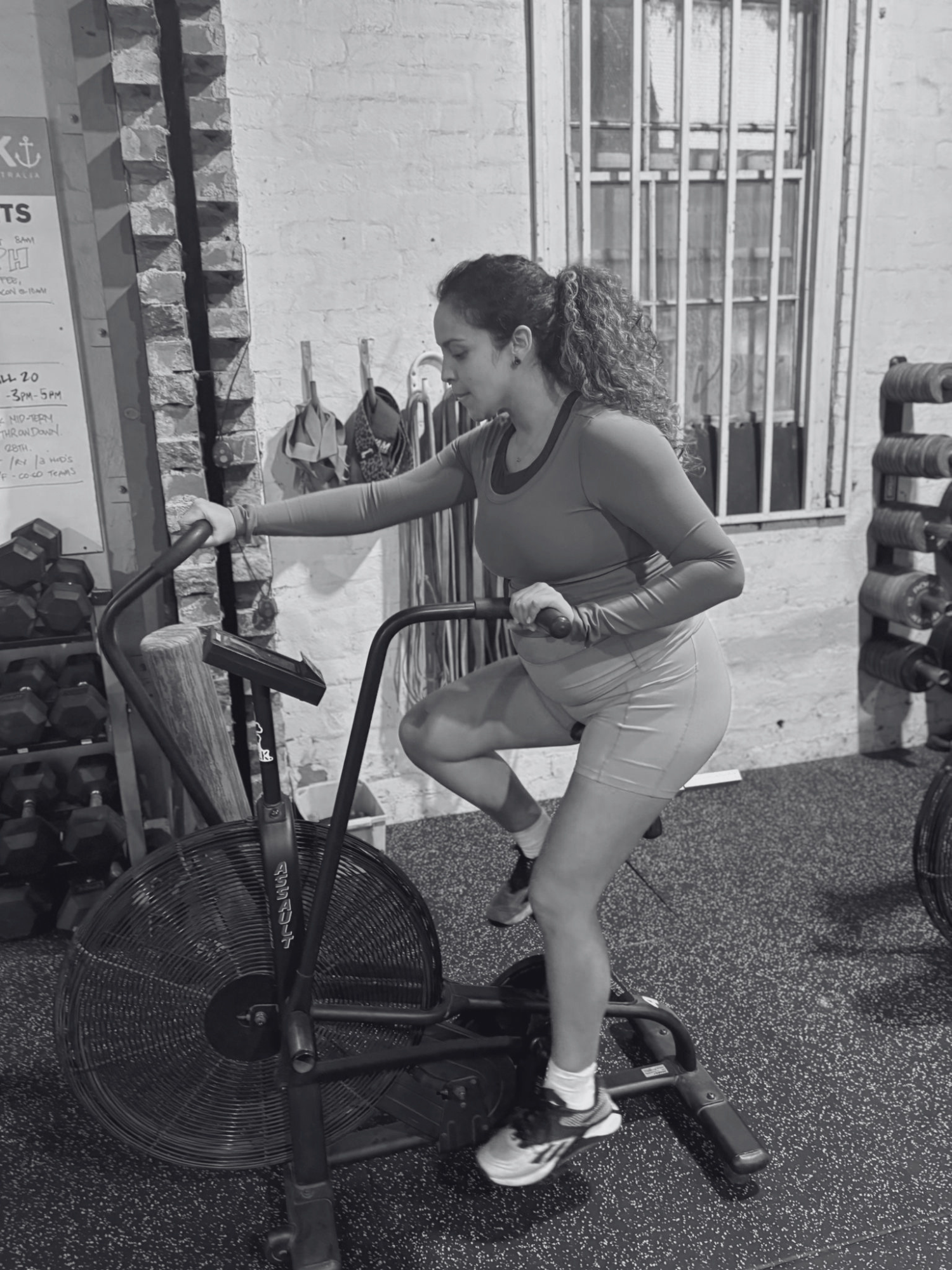 A pregnant woman with curly hair tied in a ponytail is exercising on a fan bike in a gym, wearing a fitted long-sleeve top and shorts.