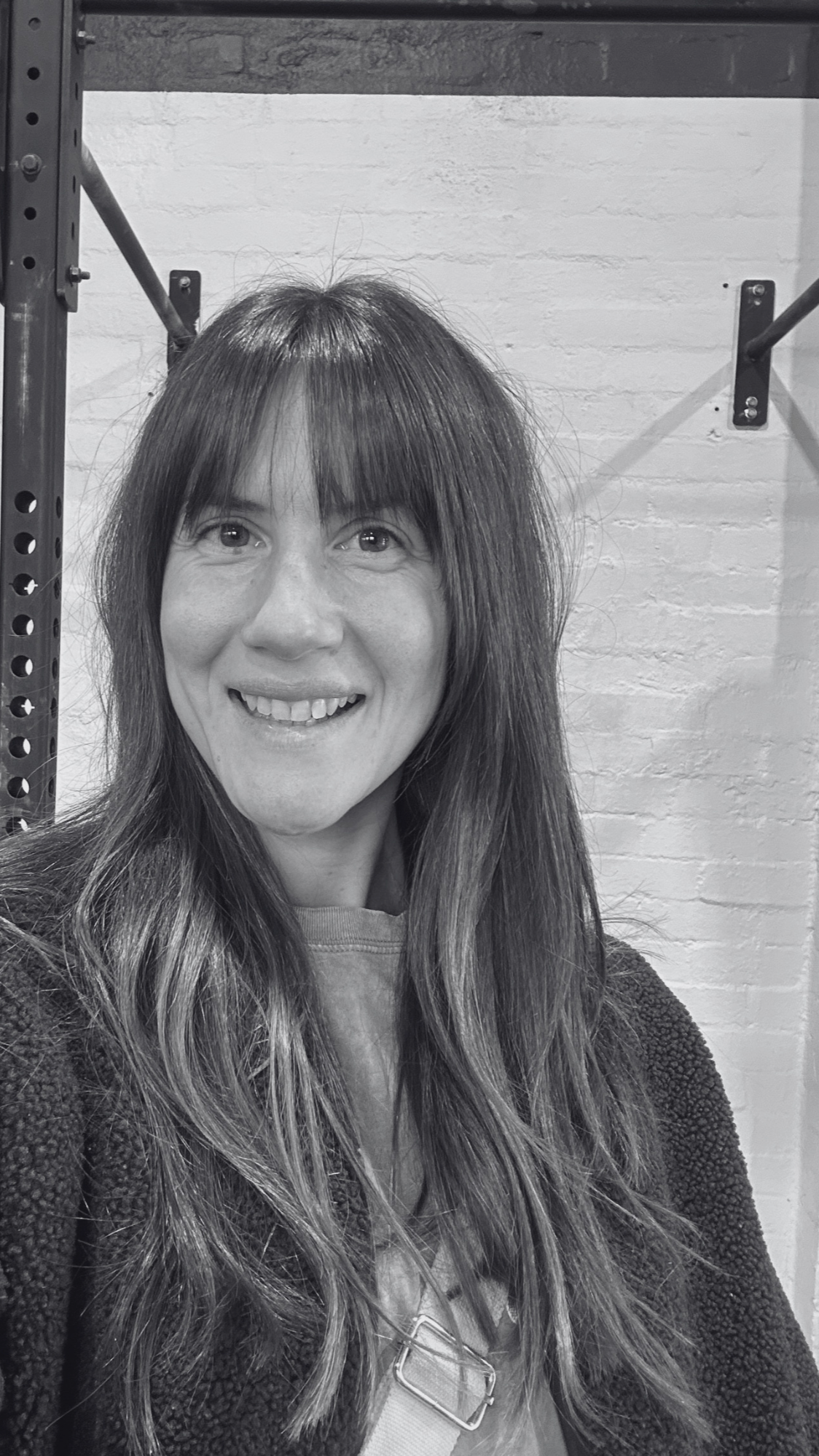 Black and white photo of a woman with long, wavy hair, smiling, standing in front of a white brick wall with hanging metal bars.