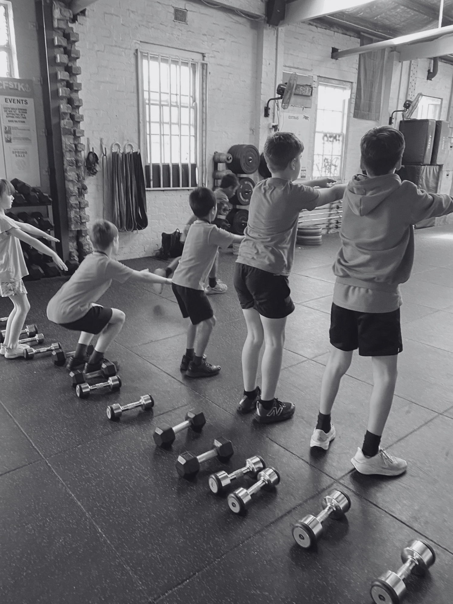 Children participating in a crossfit fitness class in a gym, performing exercises together, with dumbbells on the floor in front of them.