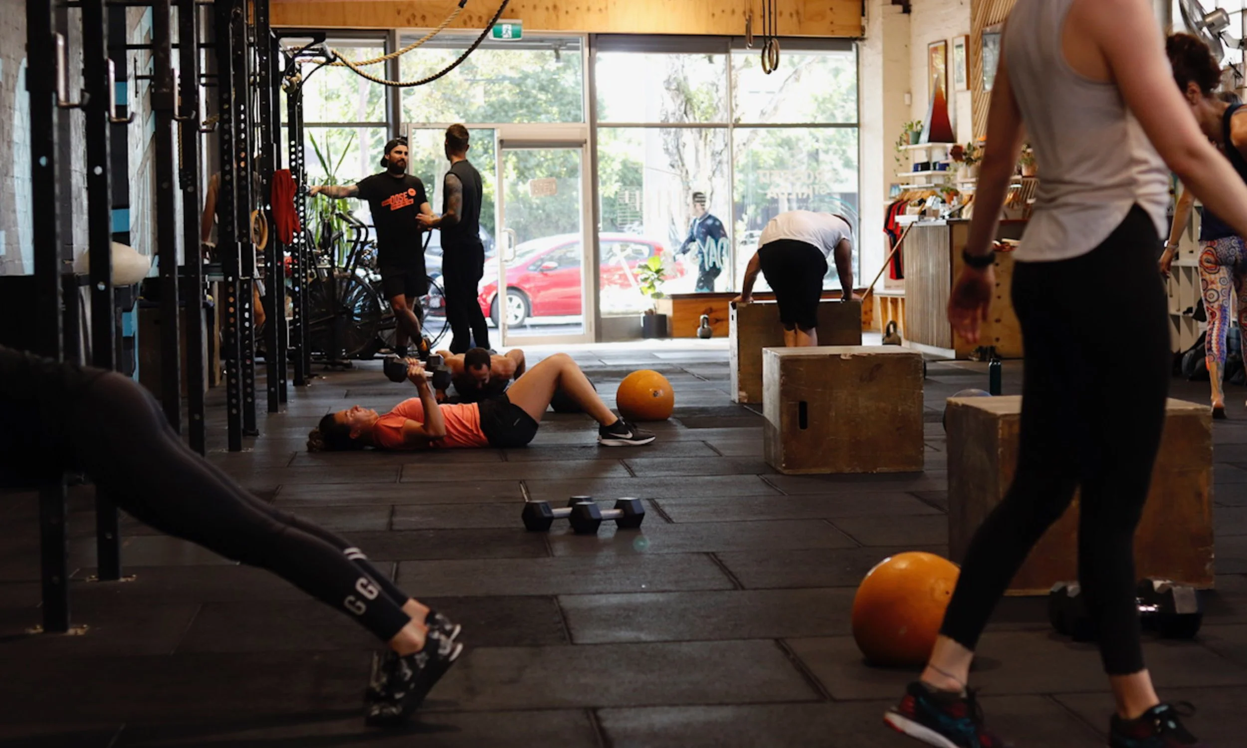 People working out in a gym with large windows, weights, and workout equipment, some engaged in exercises like lifting dumbbells and stretching.