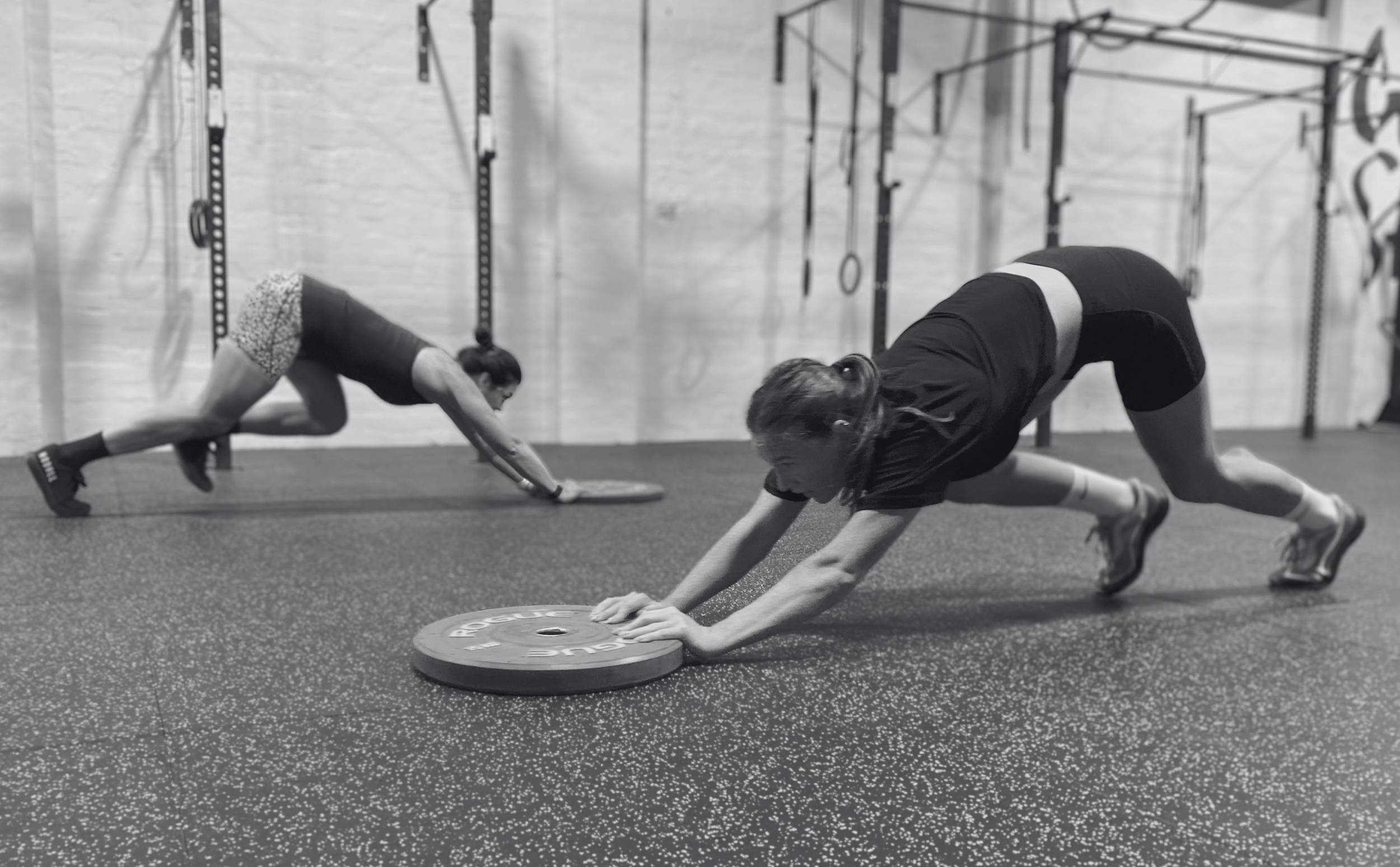 Two women in athletic clothing and sneakers performing planks on a rubber gym floor with gym equipment in the background.