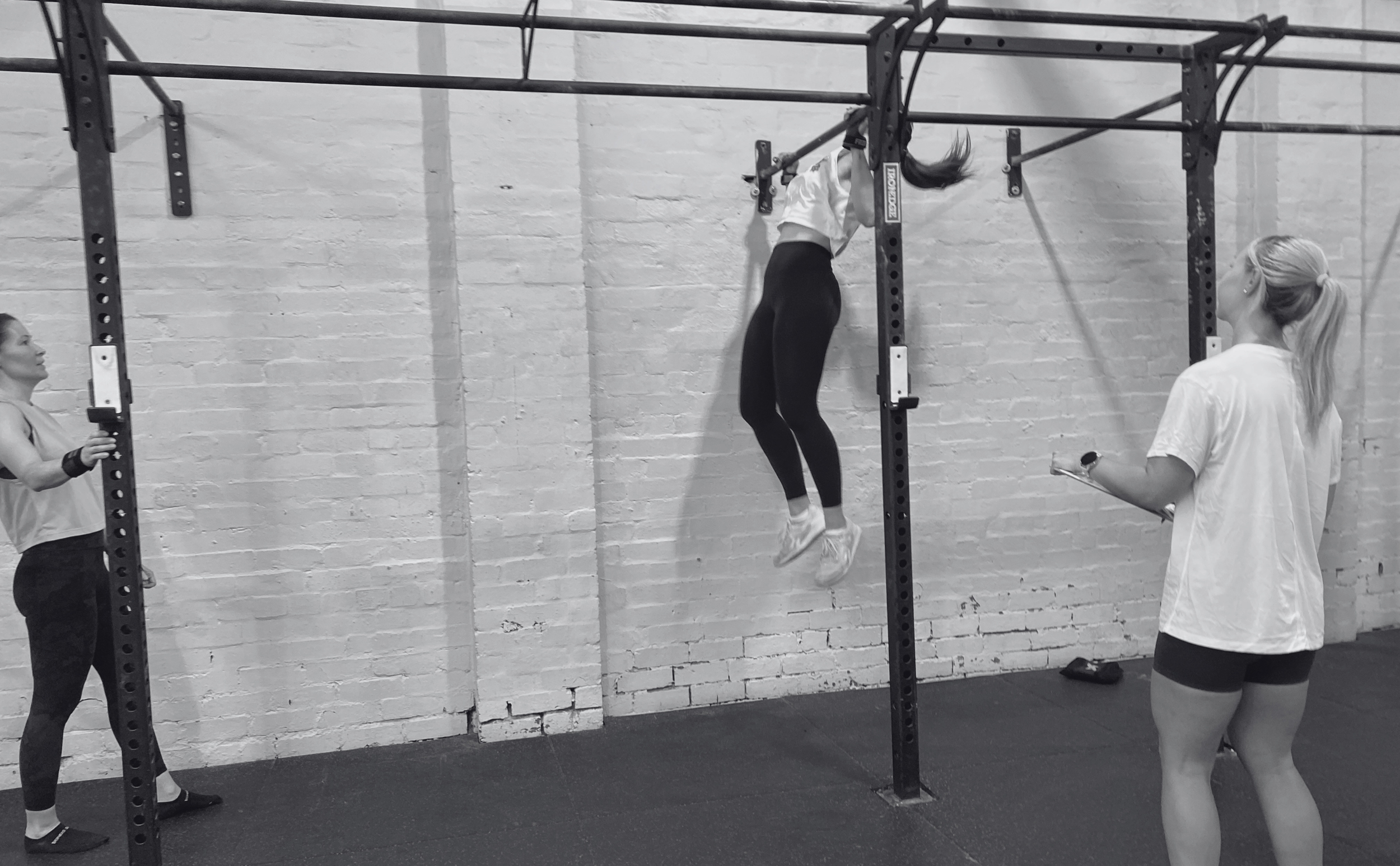 A woman is doing a pull-up on a pull-up bar in a gym, while two women observe; three women are involved in a fitness class, with an exposed brick wall background.