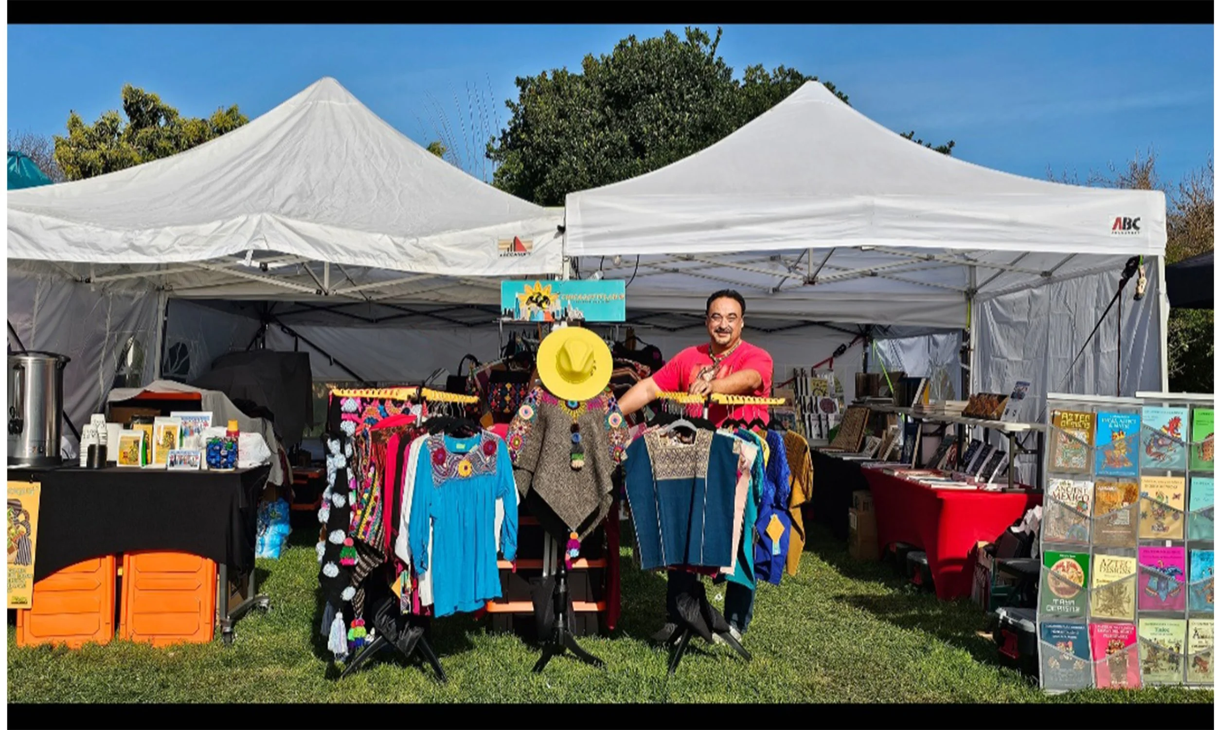A man is standing behind a display of colorful clothing at an outdoor market stall, with white tents and trees in the background under a blue sky.