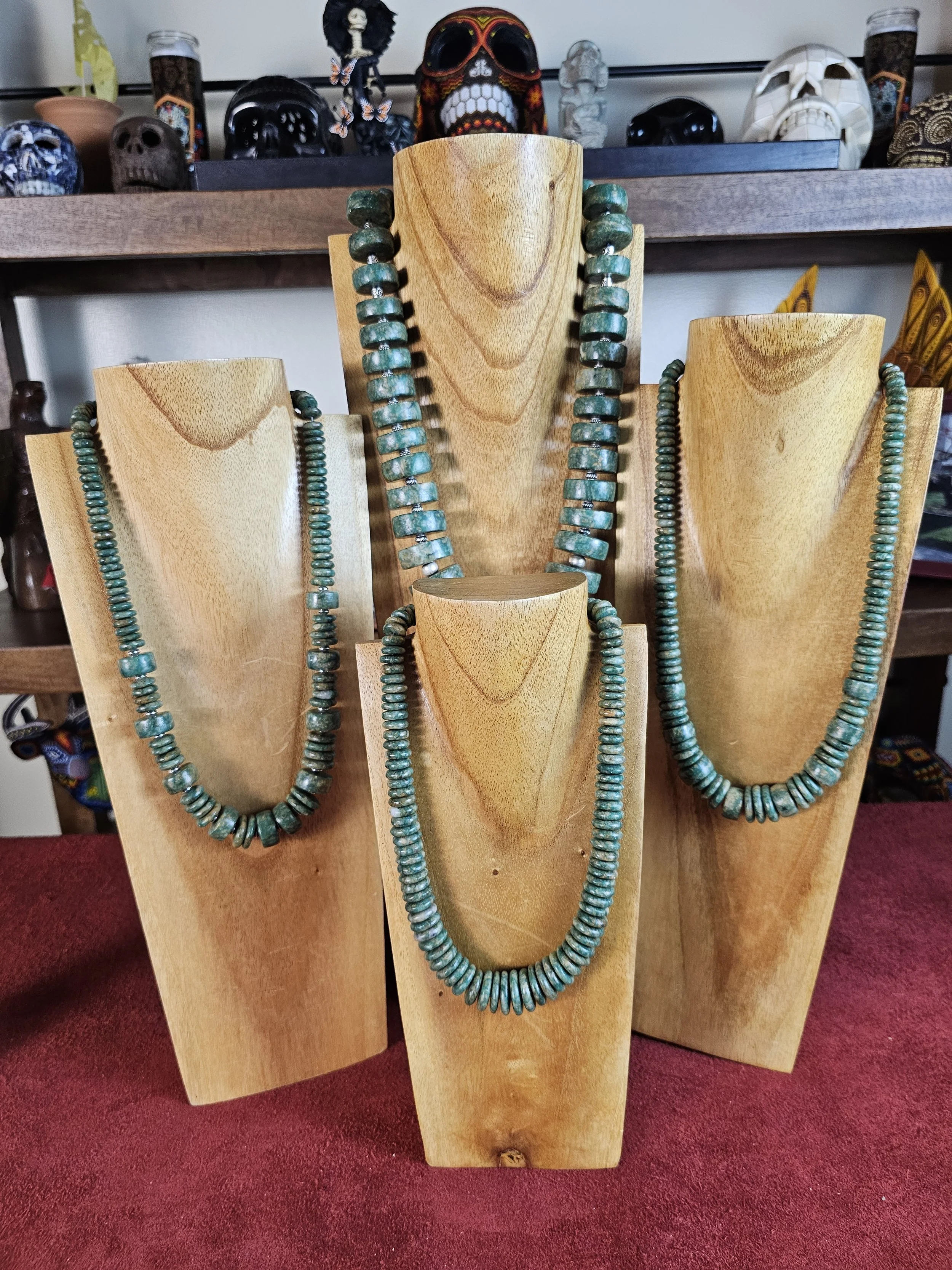 Various turquoise beaded necklaces displayed on wooden busts with a collection of skull sculptures and artifacts in the background.