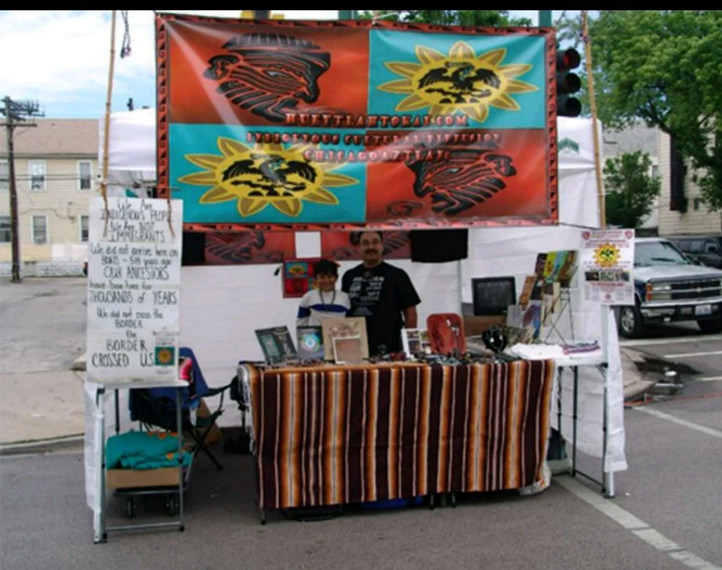 A booth at an outdoor event with a striped tablecloth and a large two-tone banner featuring a black eagle and a yellow sun with a black animal silhouette. Two people are standing behind the table, which displays artwork and merchandise, with a sign on the left expressing gratitude to veterans.