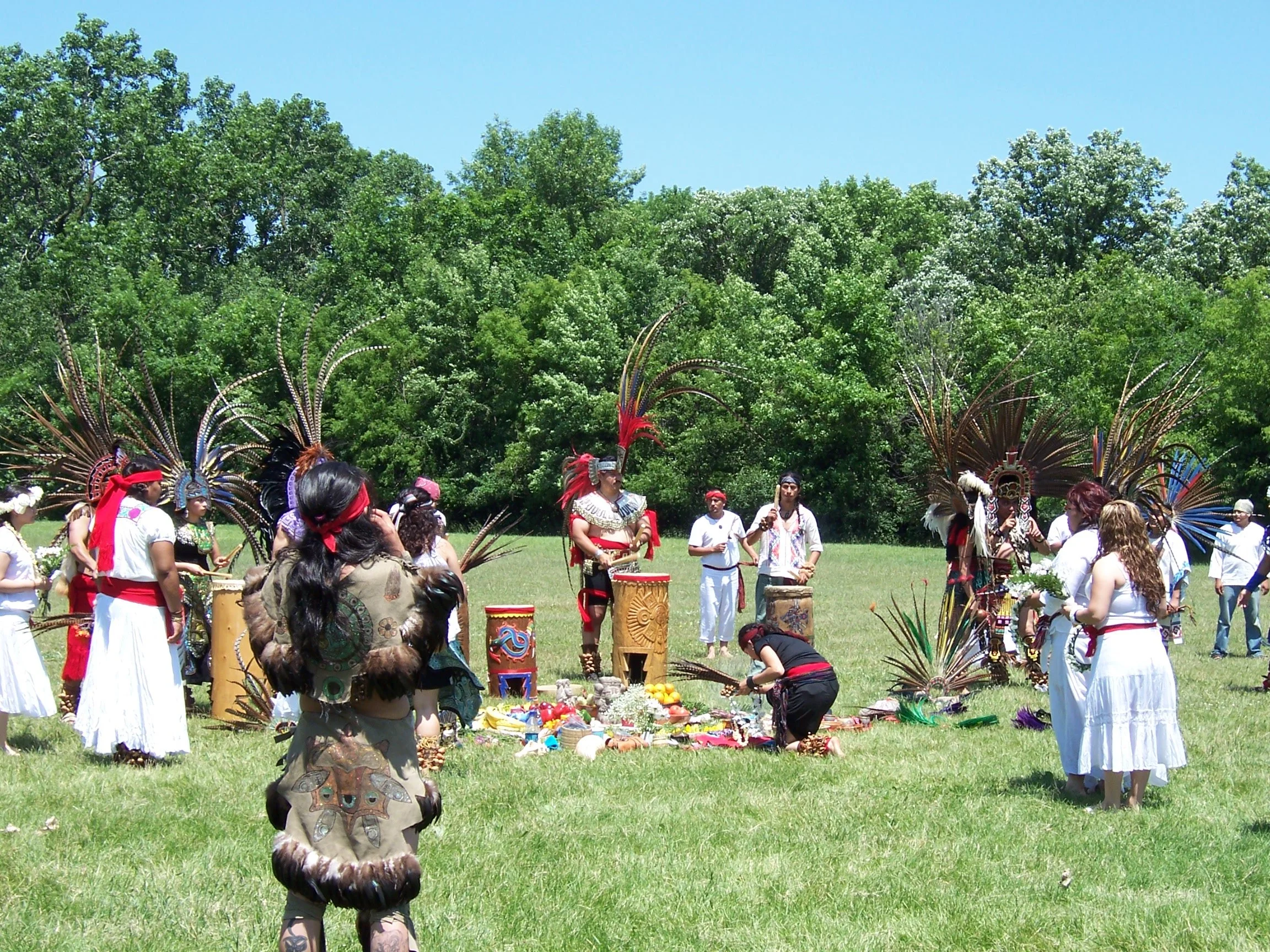 A group of Indigenous people dressed in traditional attire participating in a cultural ceremony outdoors, with trees and a clear blue sky in the background.