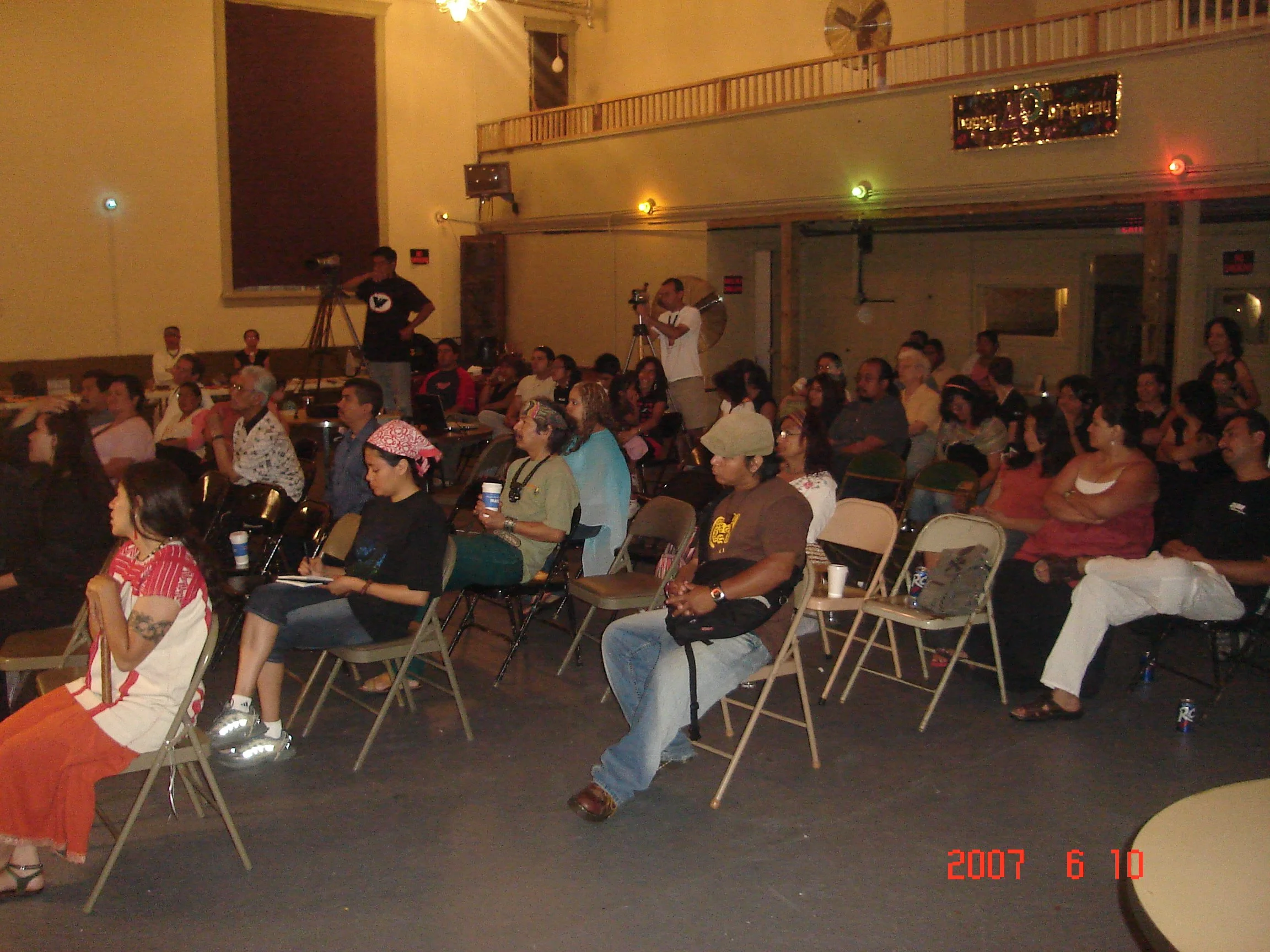 An indoor event with a large audience seated on folding chairs, some people on stage, a person with a camera, and colorful lights on the wall. The date is June 10, 2007.