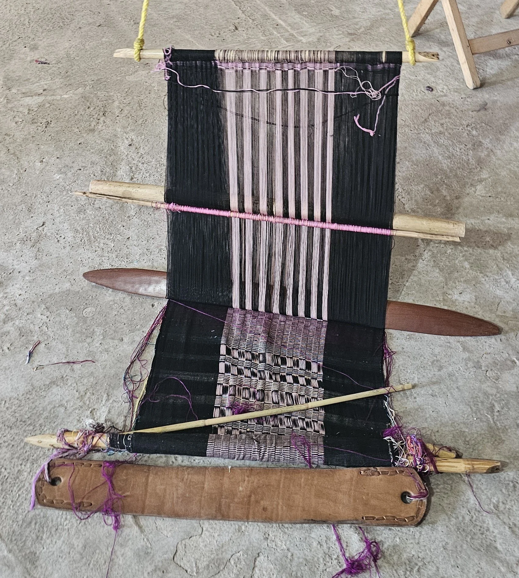 A weaving loom with black and pink threads, wooden frame, and purple yarn on a concrete floor.