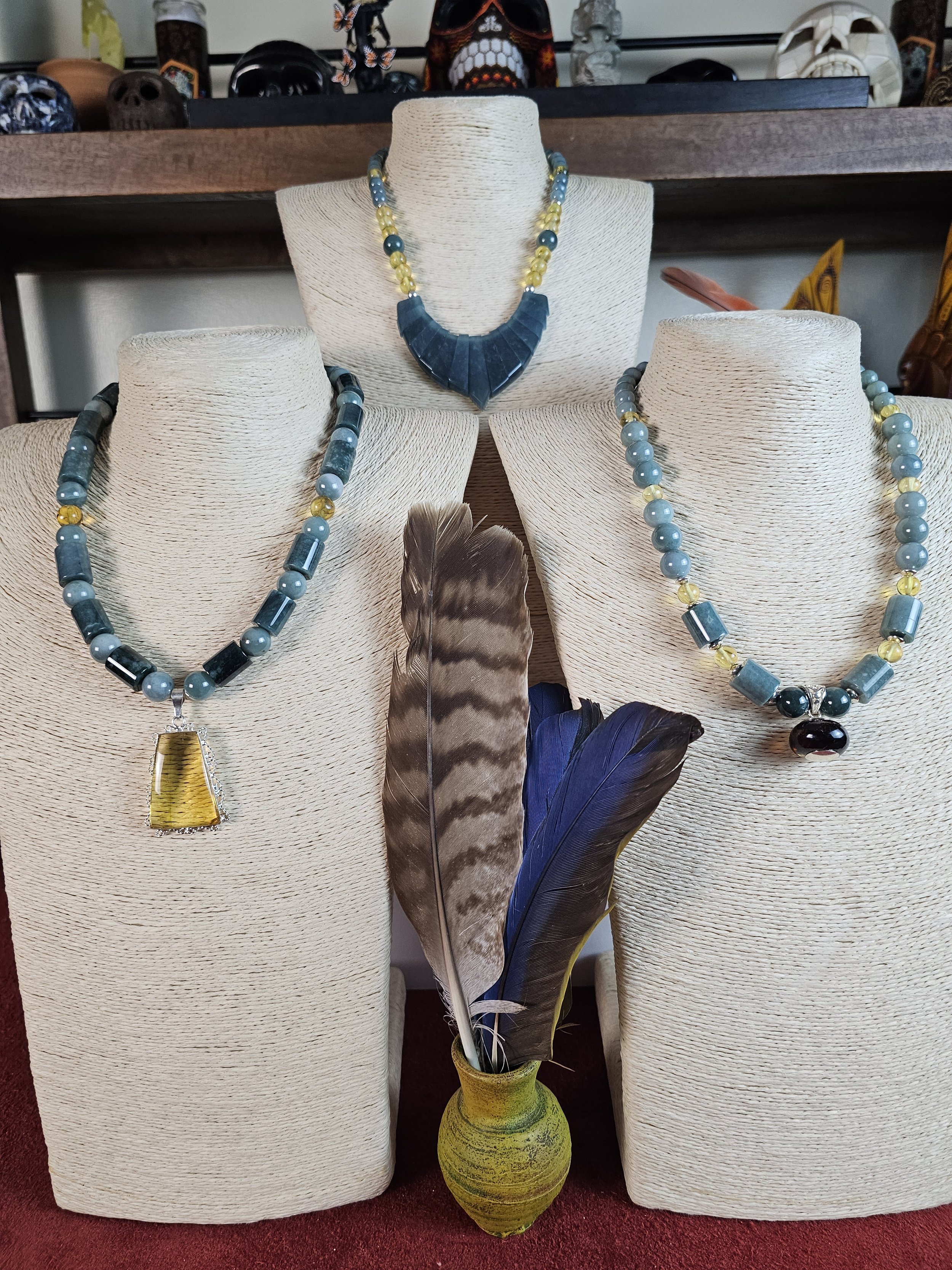 Display of three bead necklaces on beige padding bust stands, with a yellow-green vase holding large feathers placed in front.