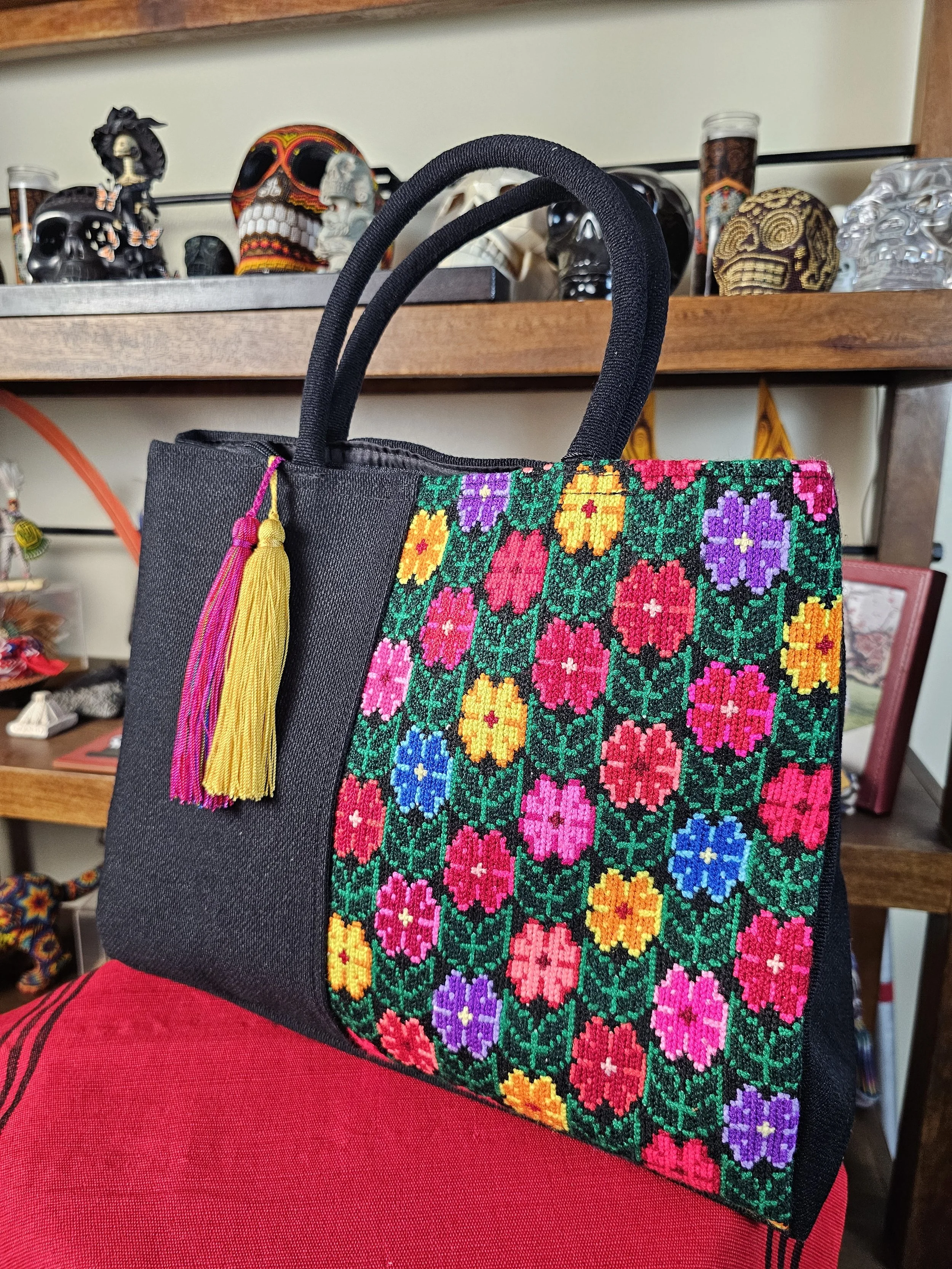 A black tote bag with a colorful embroidered floral pattern on the front, adorned with yellow and pink tassels, is resting on a person's lap. In the background, there is a wooden shelf displaying decorative skull sculptures and various small items.