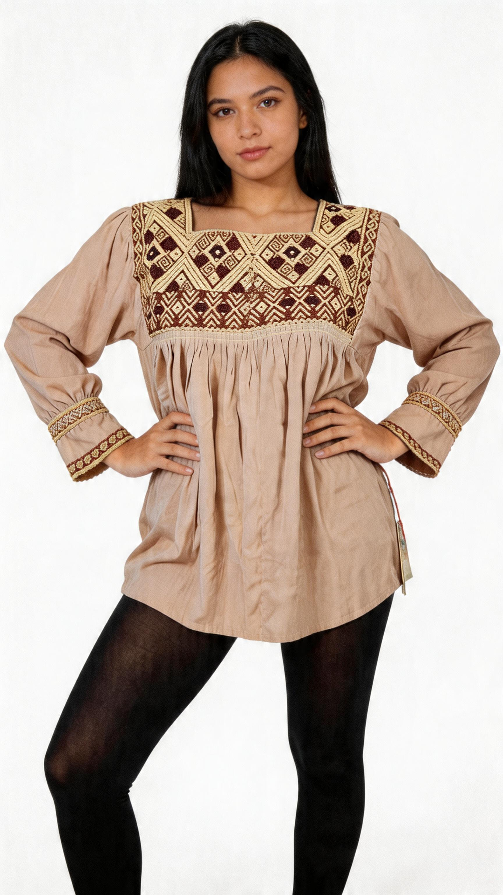 A woman wearing a beige tunic with embroidered details on the chest and sleeves, black tights, and posing with hands on hips in front of a plain white background.