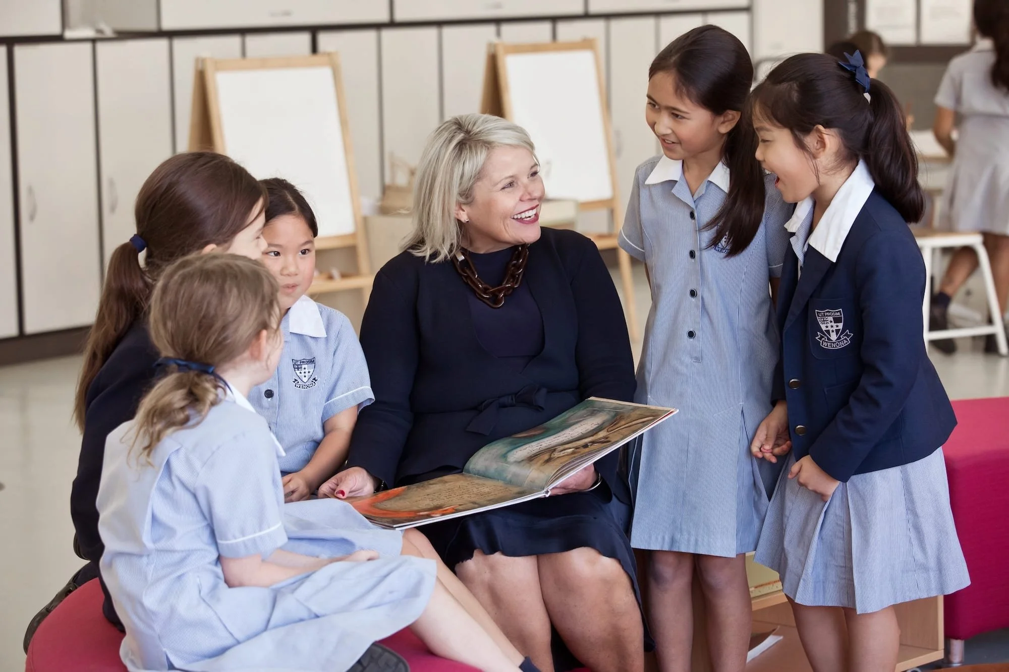 A group of young girls in school uniforms gathered around a woman with short blonde hair who is holding an illustrated book. They appear to be in a classroom setting, engaging with the woman as she shares a story or information.