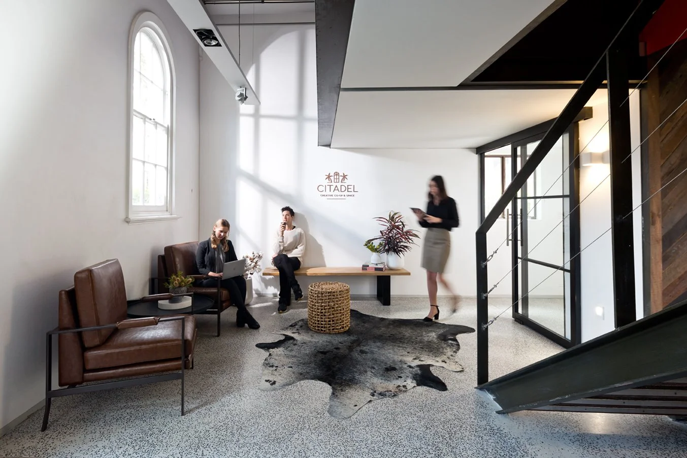 Modern office lobby with white walls, large arched window, leather chairs, a bench, plants, a cowhide rug, and people using laptops and smartphones