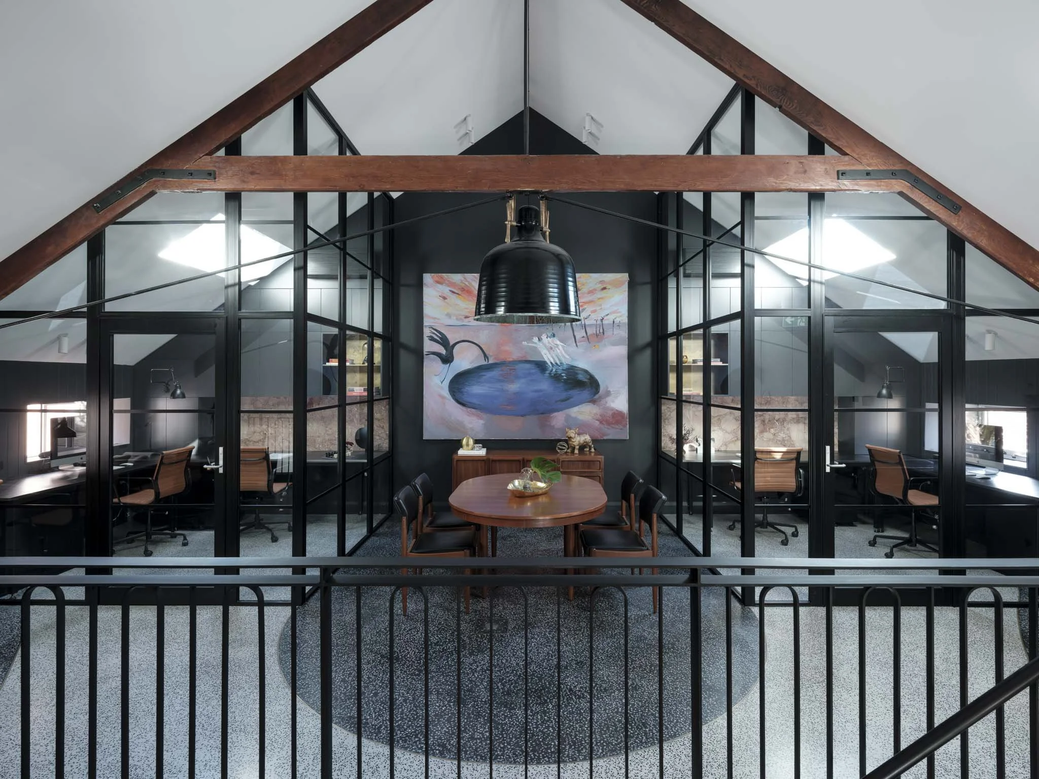 Interior view of an office space with a glass wall, a black pendant light, a wooden table, and chairs. A large abstract painting is on the back wall.