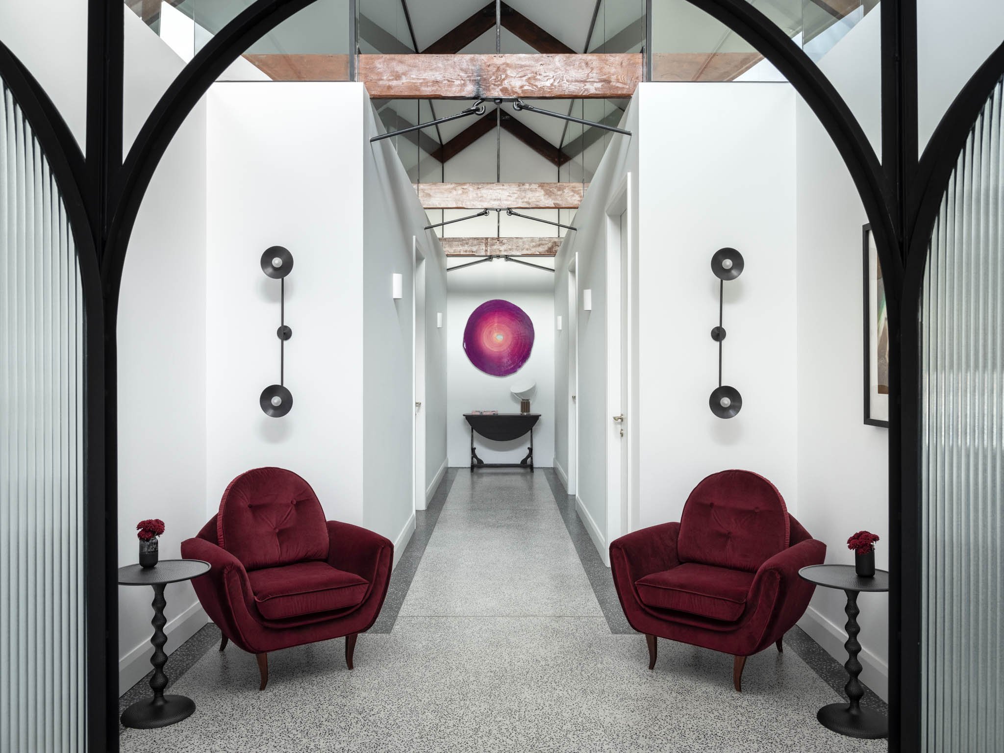A hallway with two red velvet armchairs and small side tables with flowers, framed by black arched structures, leading to a wall with a pink and purple circular artwork.