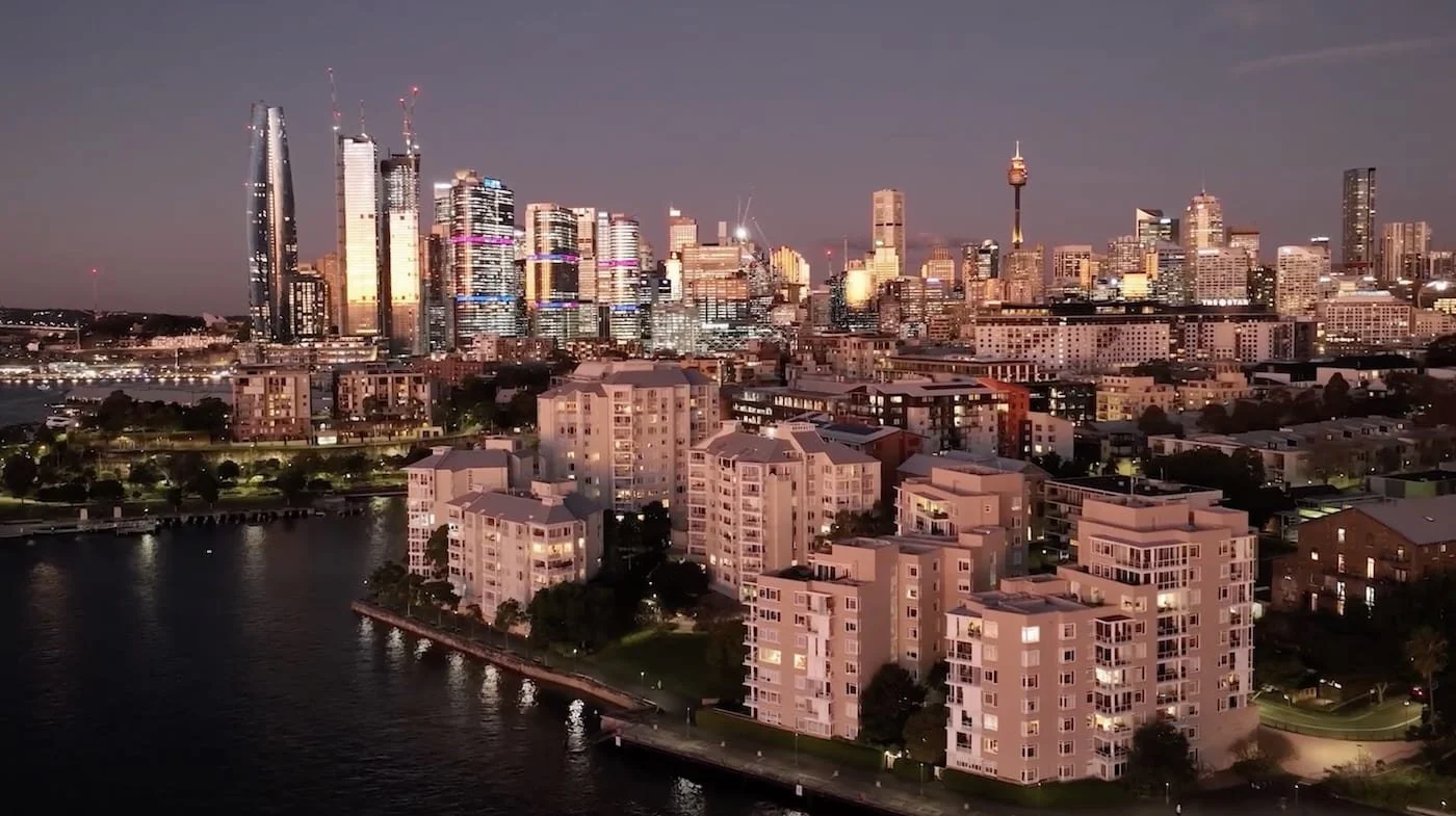 City skyline at dusk with modern skyscrapers in the background and residential buildings near a waterway in the foreground.
