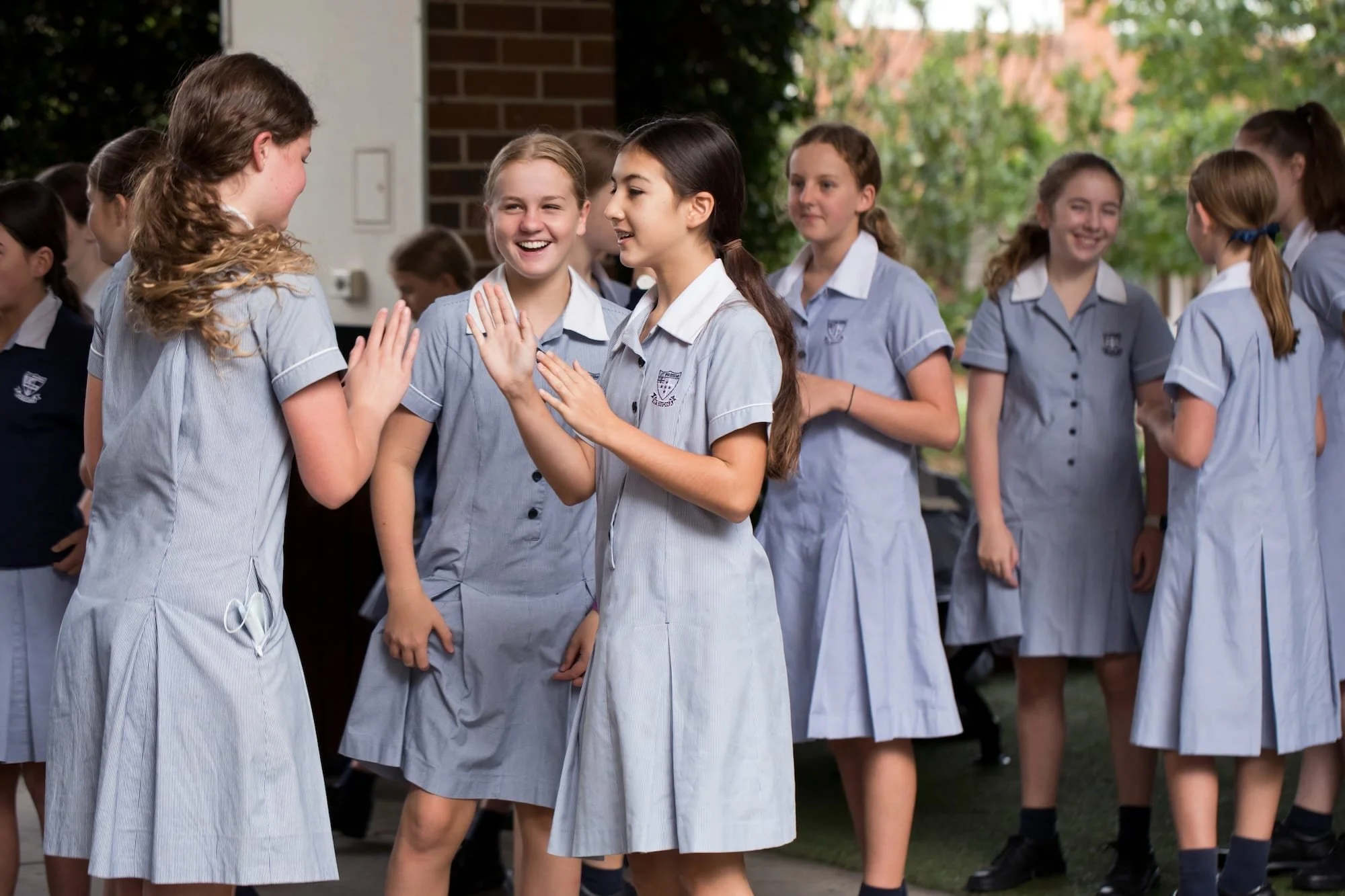 Group of schoolgirls dressed in uniforms, standing outside on a schoolyard, engaging in conversation and smiling.